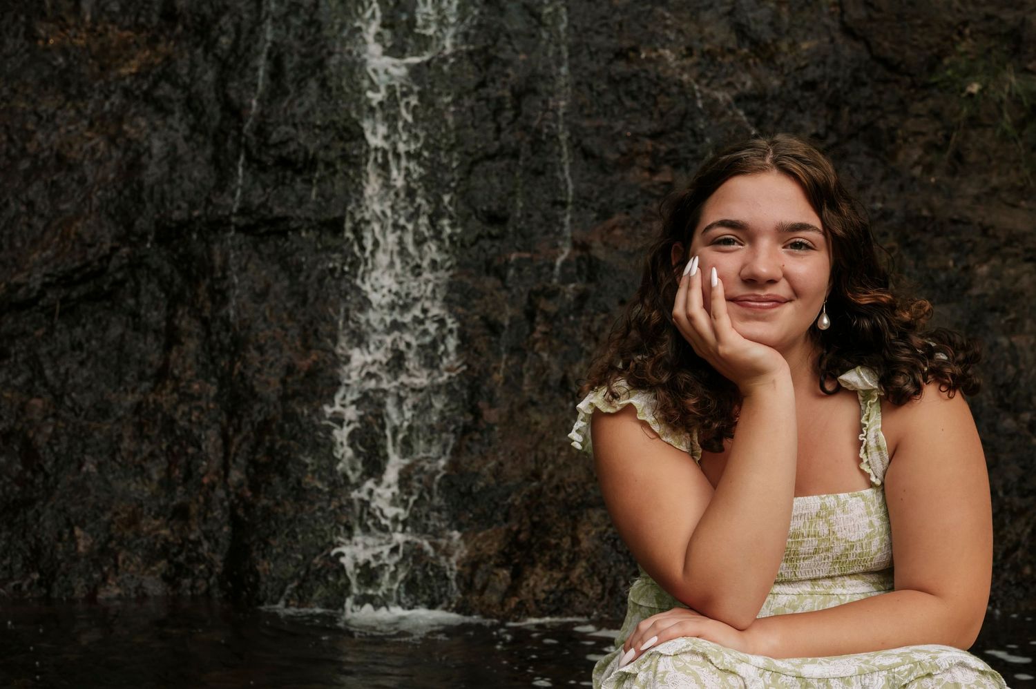 A high school senior sitting in front of a waterfall, resting her face on her hand