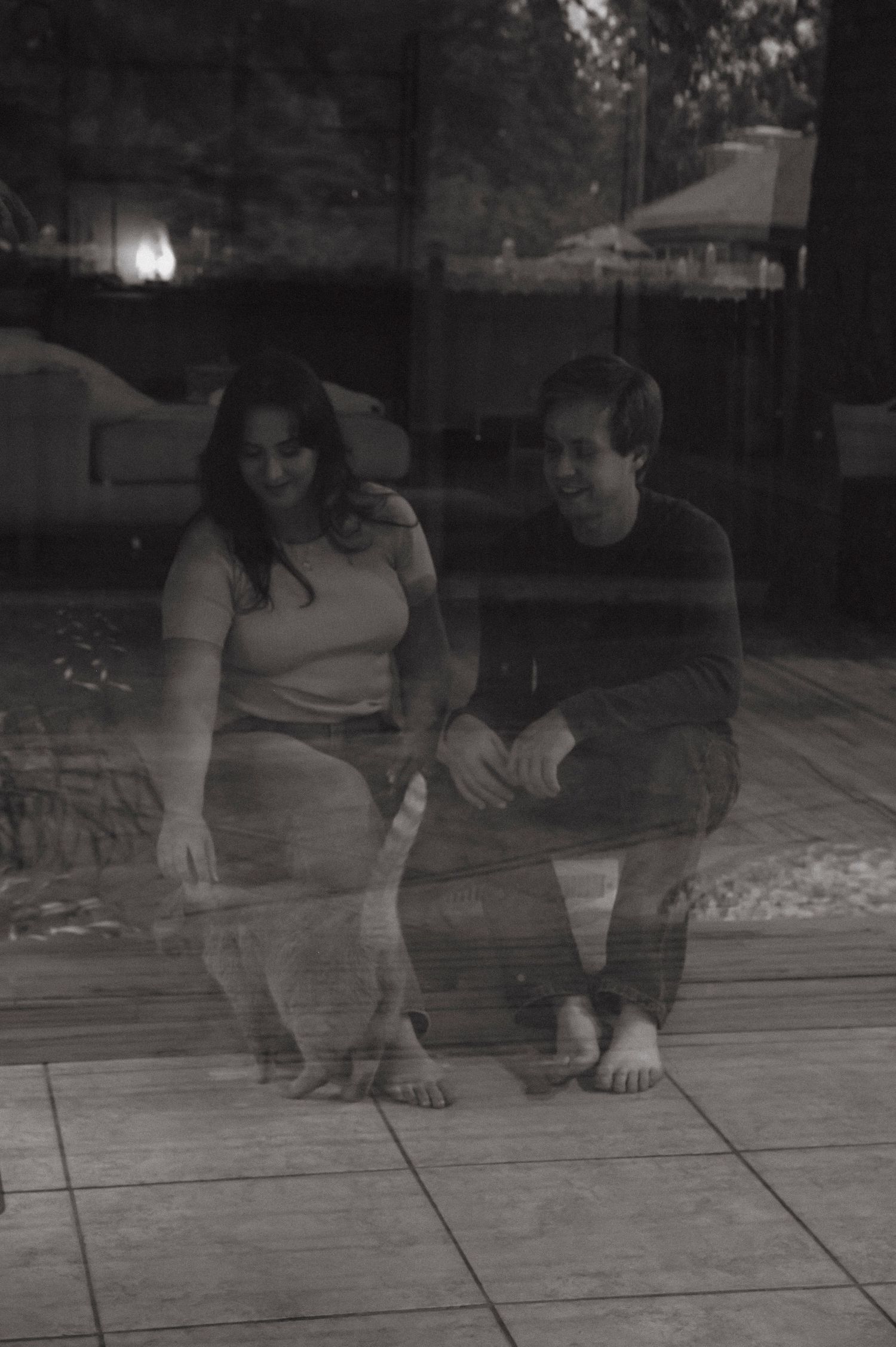 A couple sits in their home petting their cat. The photo is taken from outside through the window