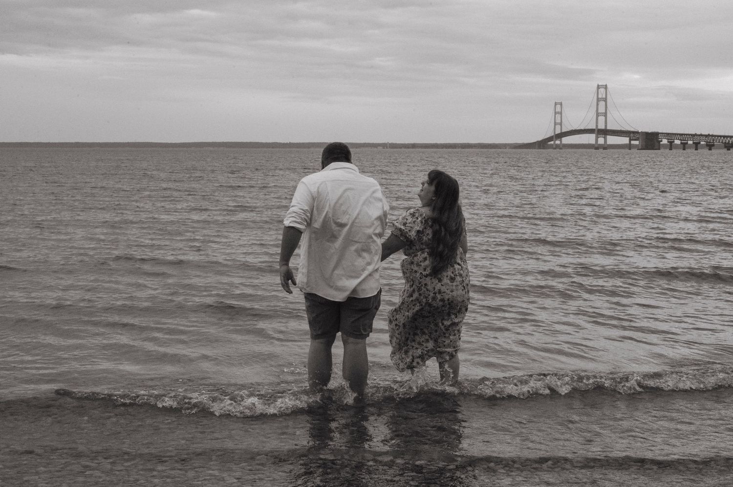 A couple runs into the water from the beach with the Mackinaw Bridge in front of them