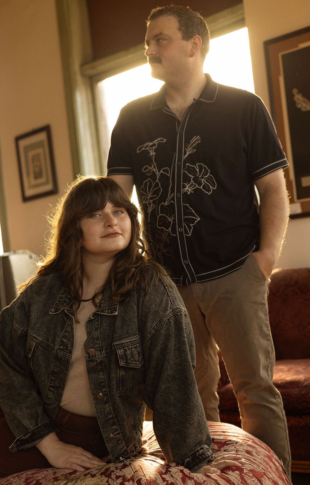 A couple, the woman sitting and the man standing behind her, in front of a large window with the sunlight illuminating them