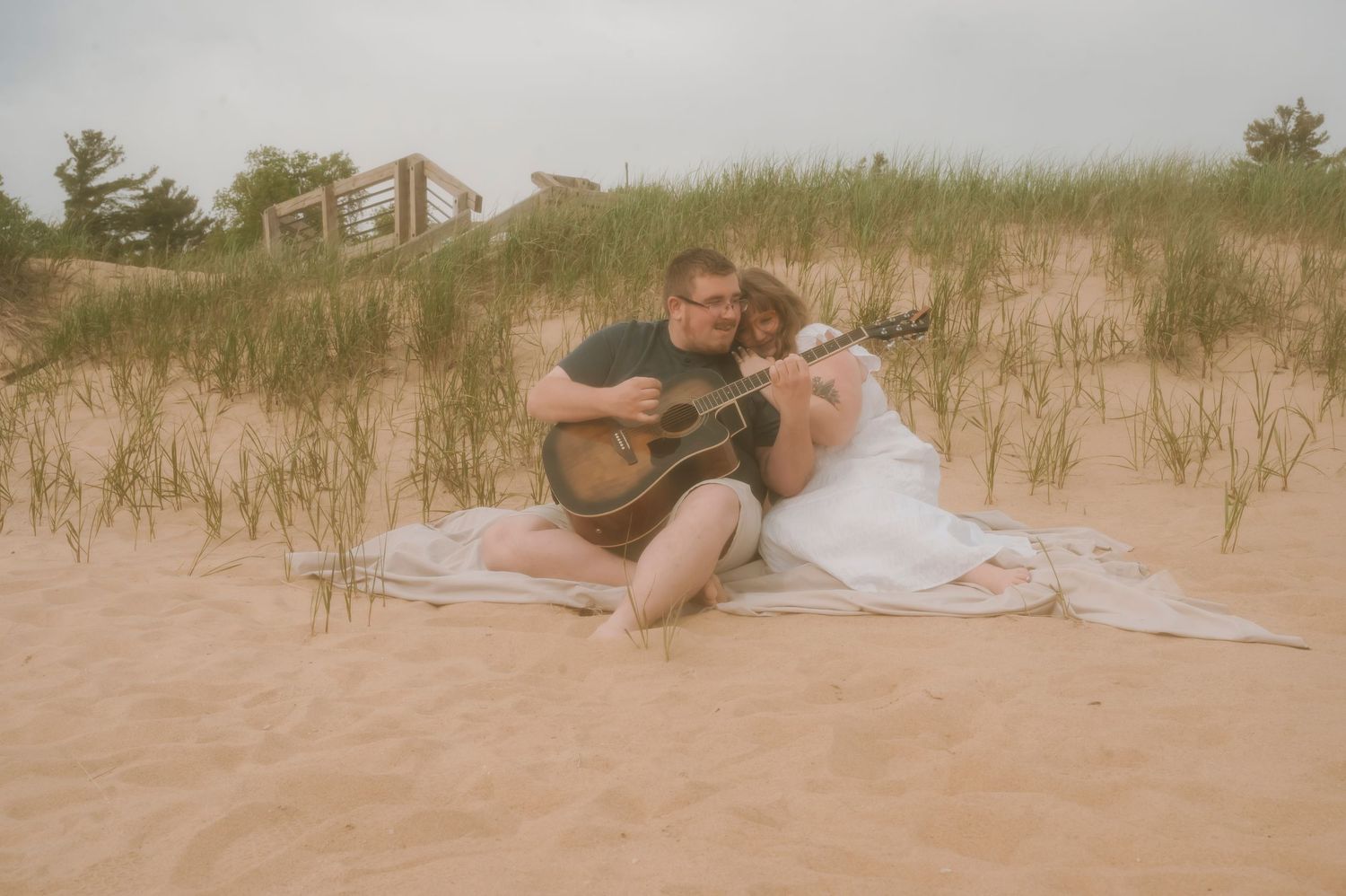 A couple on the beach, sitting on a sheet while one plays the guitar as the other snuggles up close