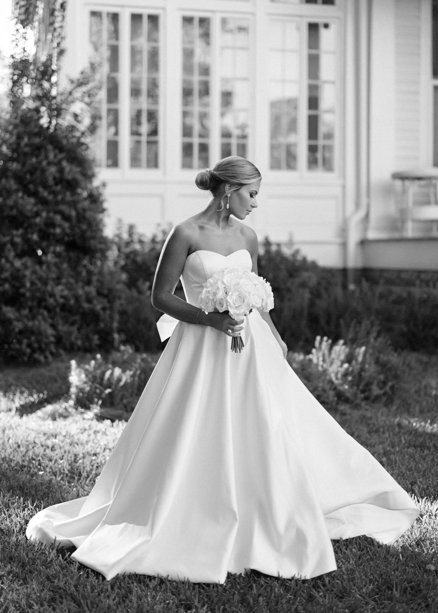 A bride in a flowing white wedding gown twirls gracefully in a garden setting, captured in black and white photography.