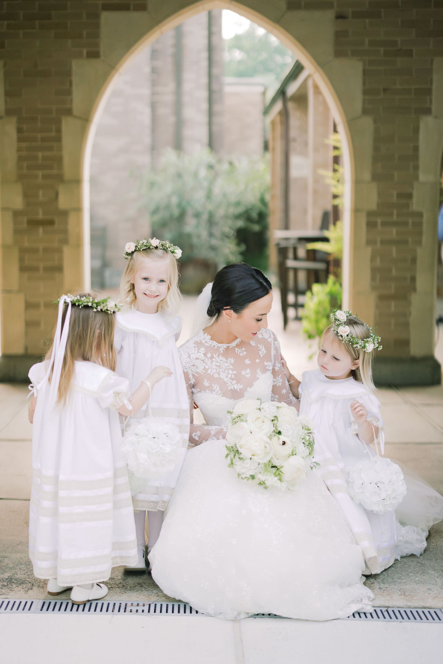 Wedding scene with bride in lace gown surrounded by flower girls in white dresses.