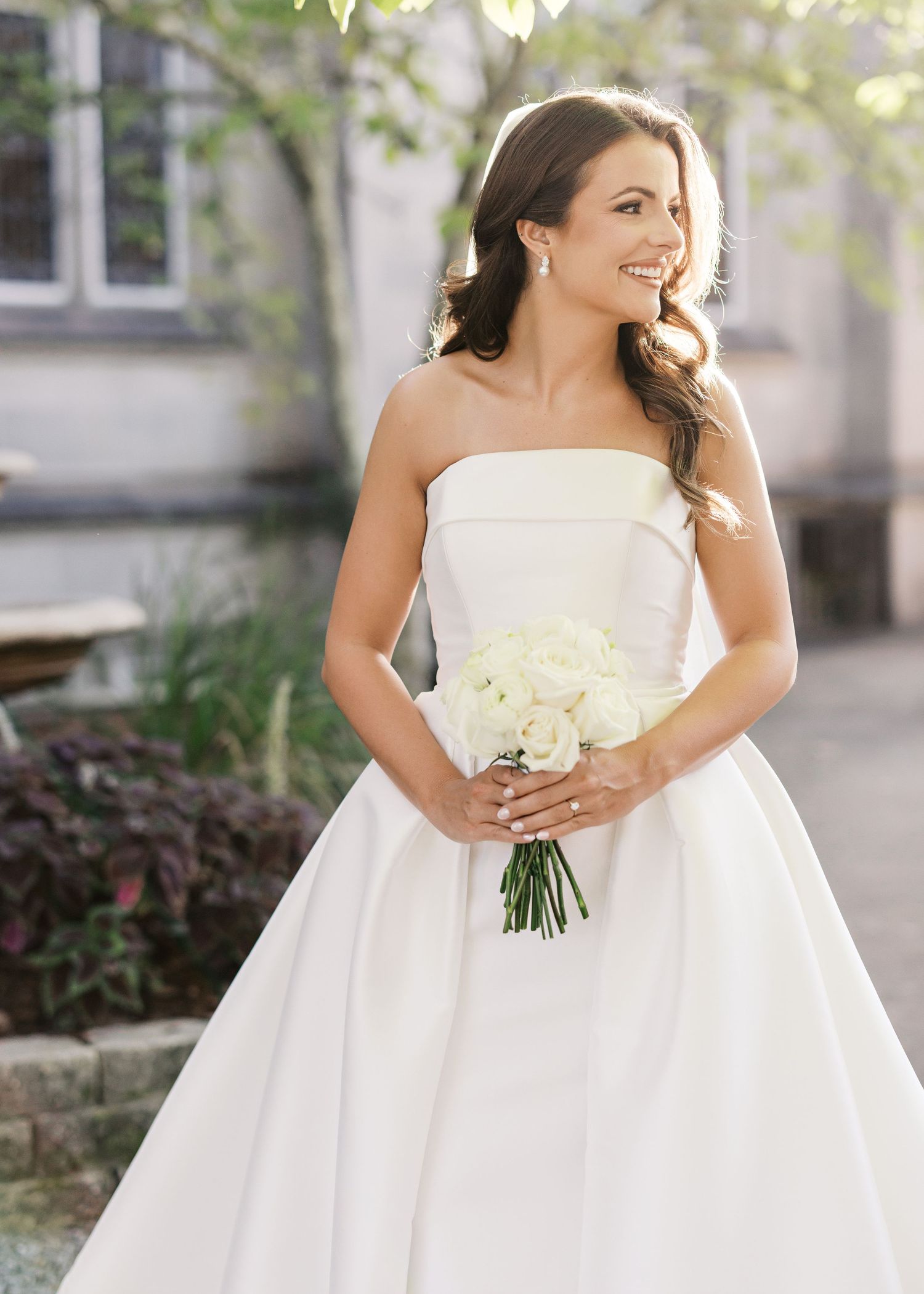 A bride in a white strapless ballgown holds a white floral bouquet while posing in a garden setting.