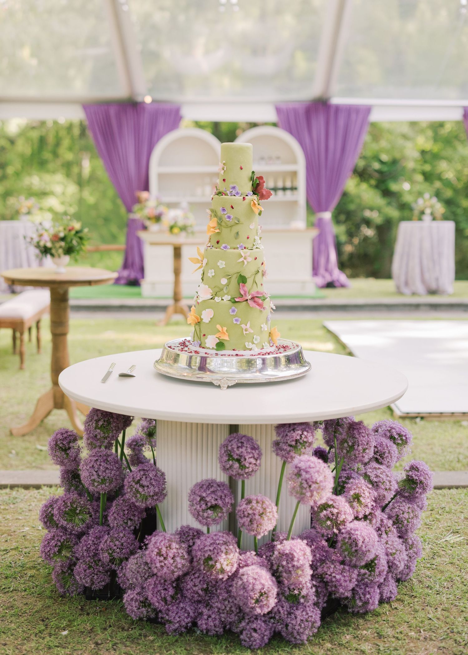 Elegant multi-tier green wedding cake adorned with flowers displayed on white table surrounded by purple allium blooms outdoors.