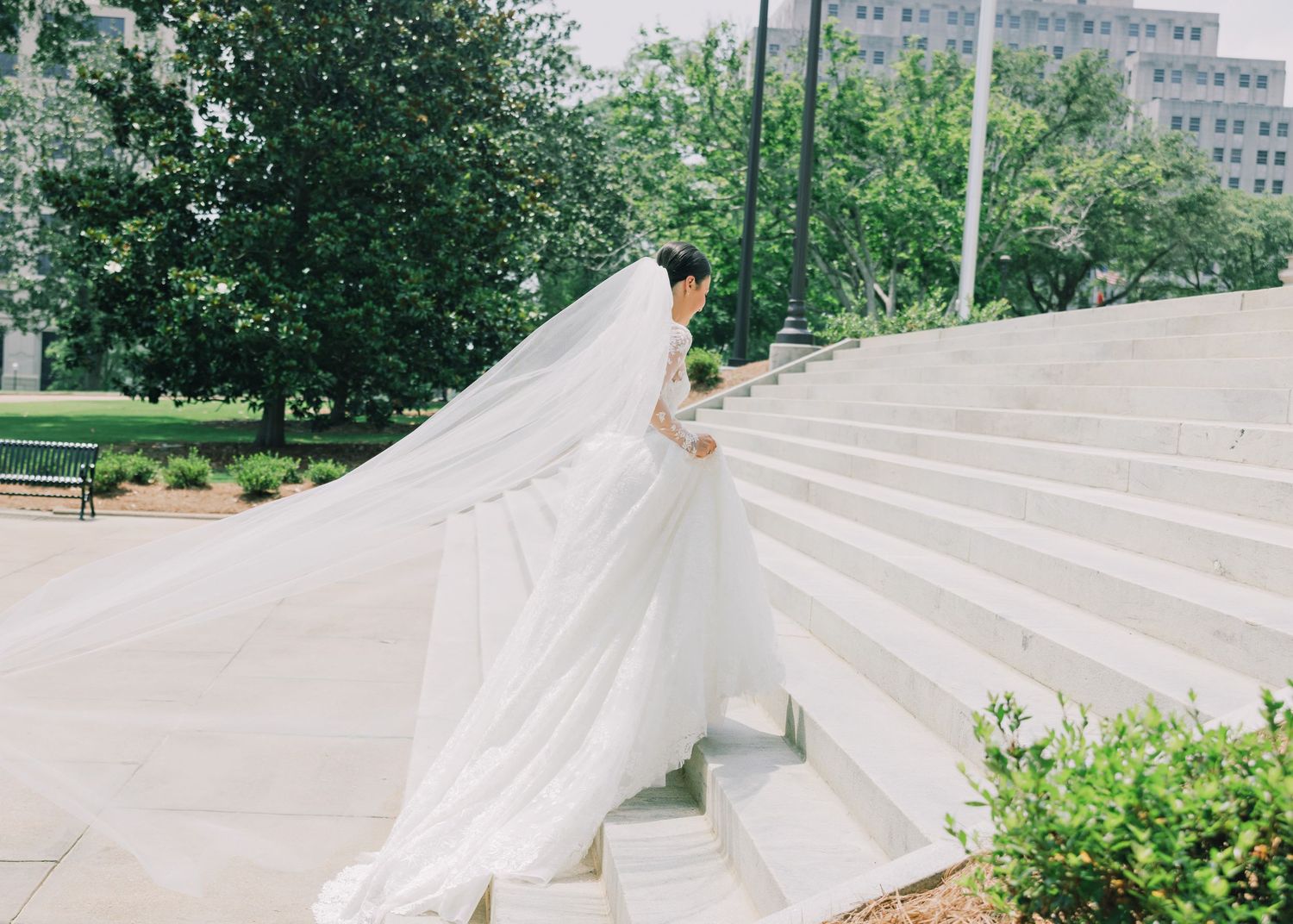 A bride in a flowing white dress ascends grand stone stairs outdoors.