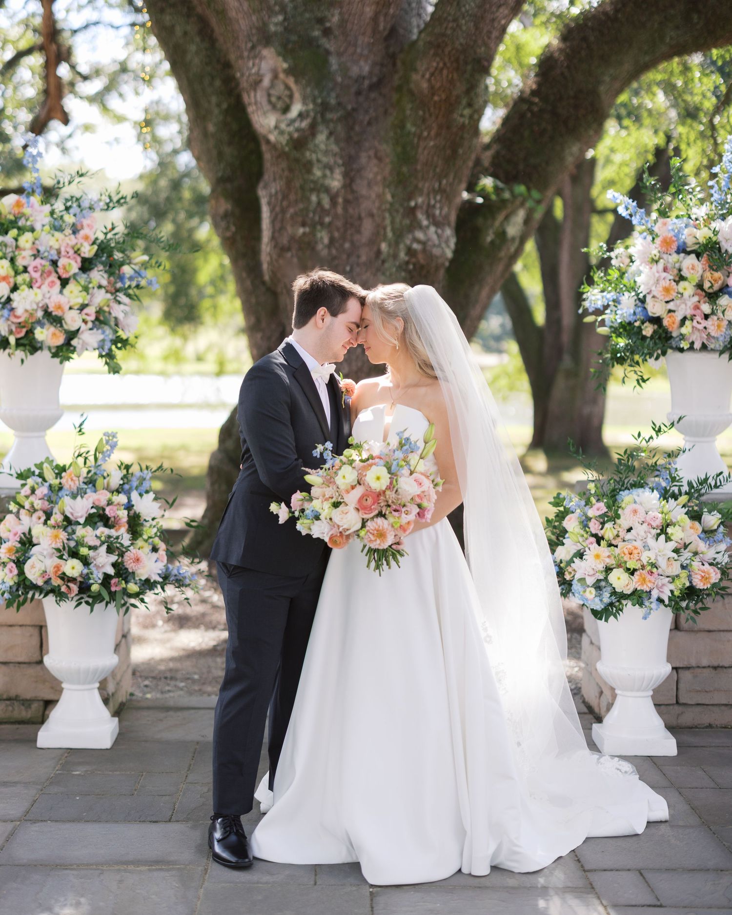 Elegant outdoor wedding ceremony setup with floral arrangements in white urns beneath large oak trees.