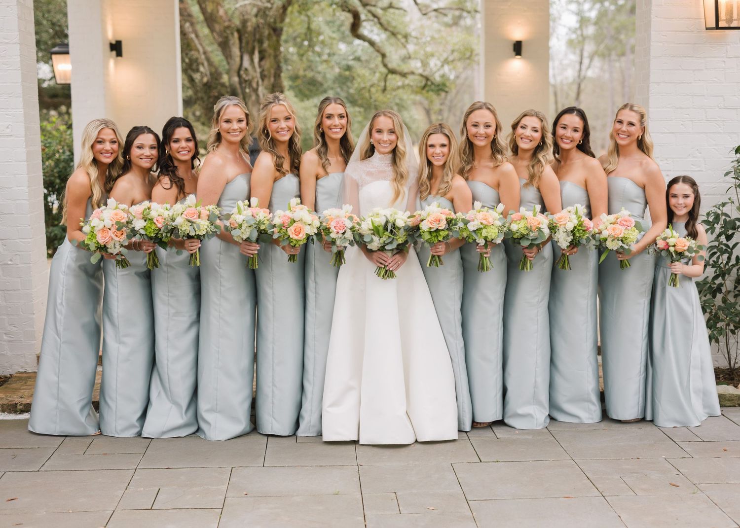 Bridesmaids in light blue dresses stand in a row with bouquets on a stone patio during an elegant outdoor wedding.