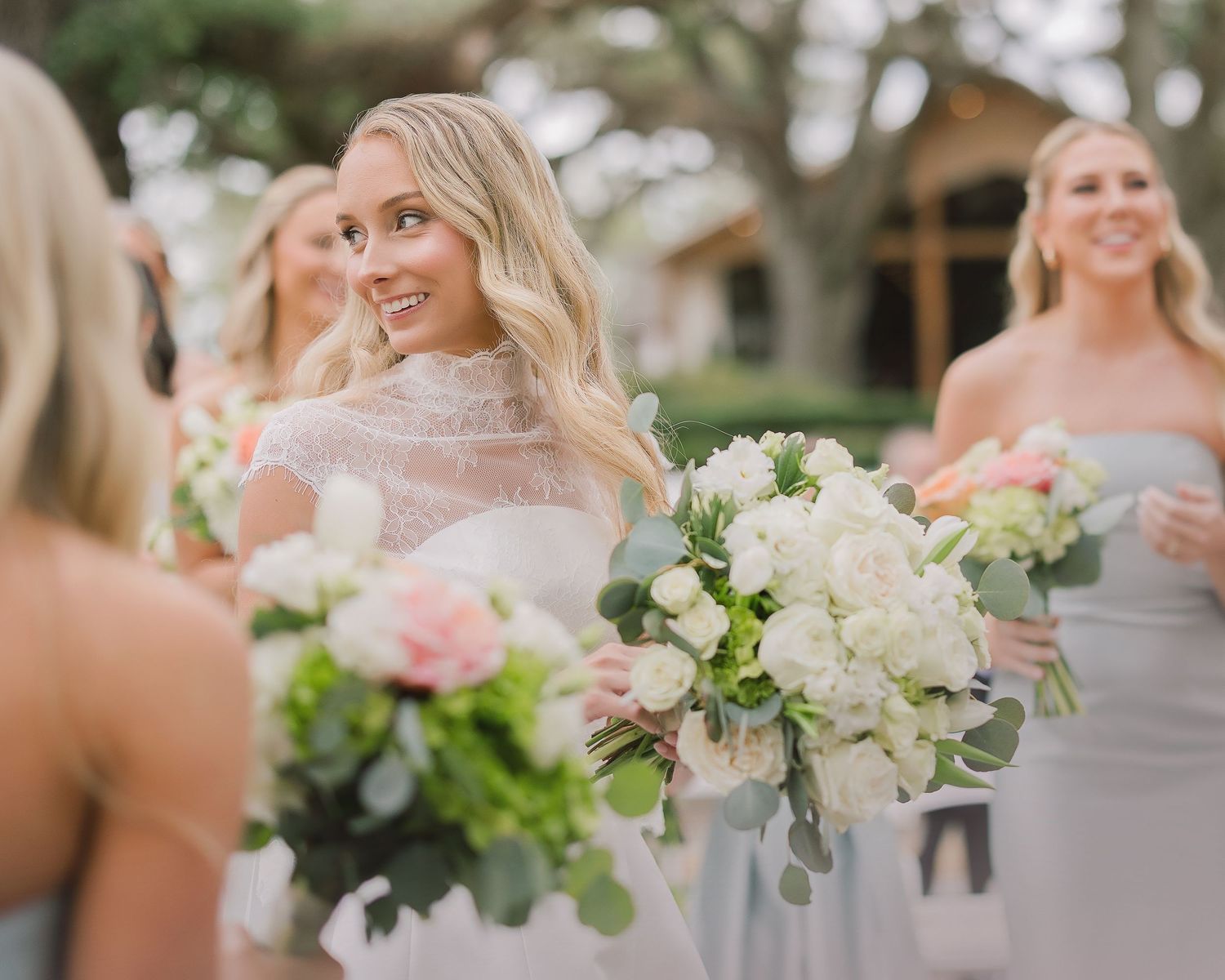 Bridesmaids in light gray dresses hold elegant bouquets of white and pink flowers at an outdoor wedding celebration.