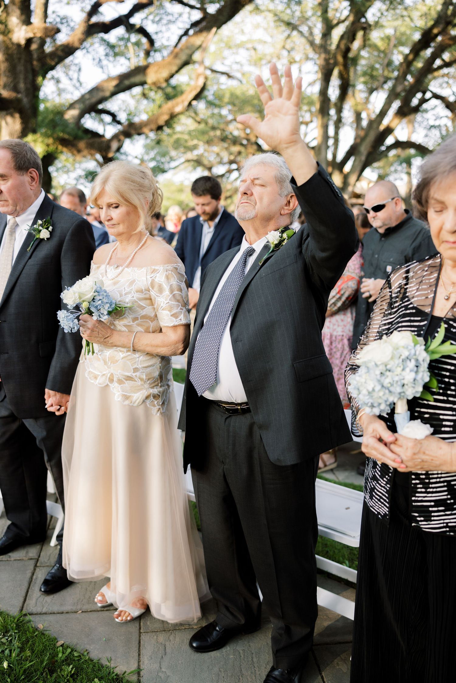 Wedding guests in formal attire holding bouquets gather outdoors beneath oak trees.