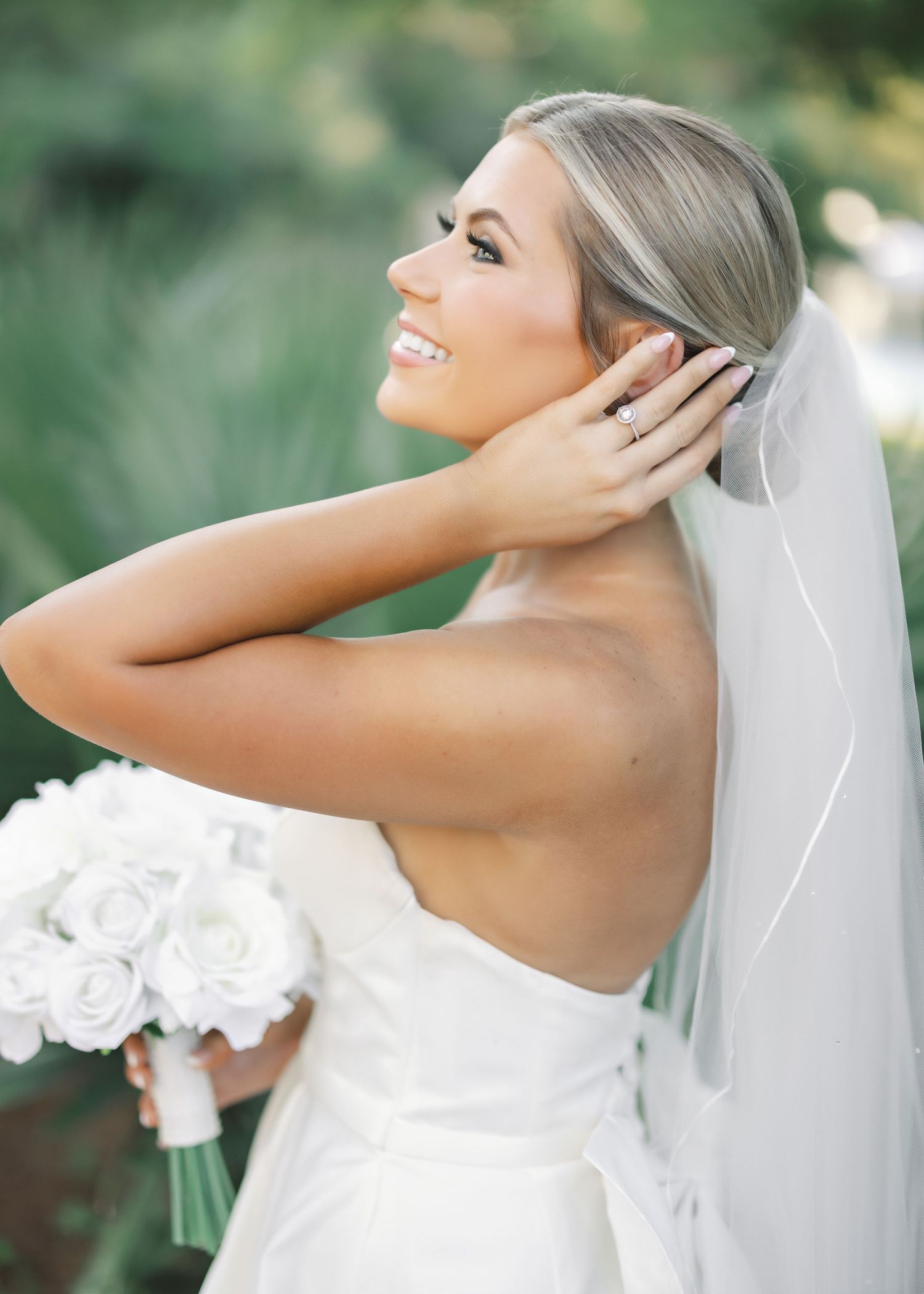 Bride in strapless white gown and veil poses with white rose bouquet in an outdoor garden setting.