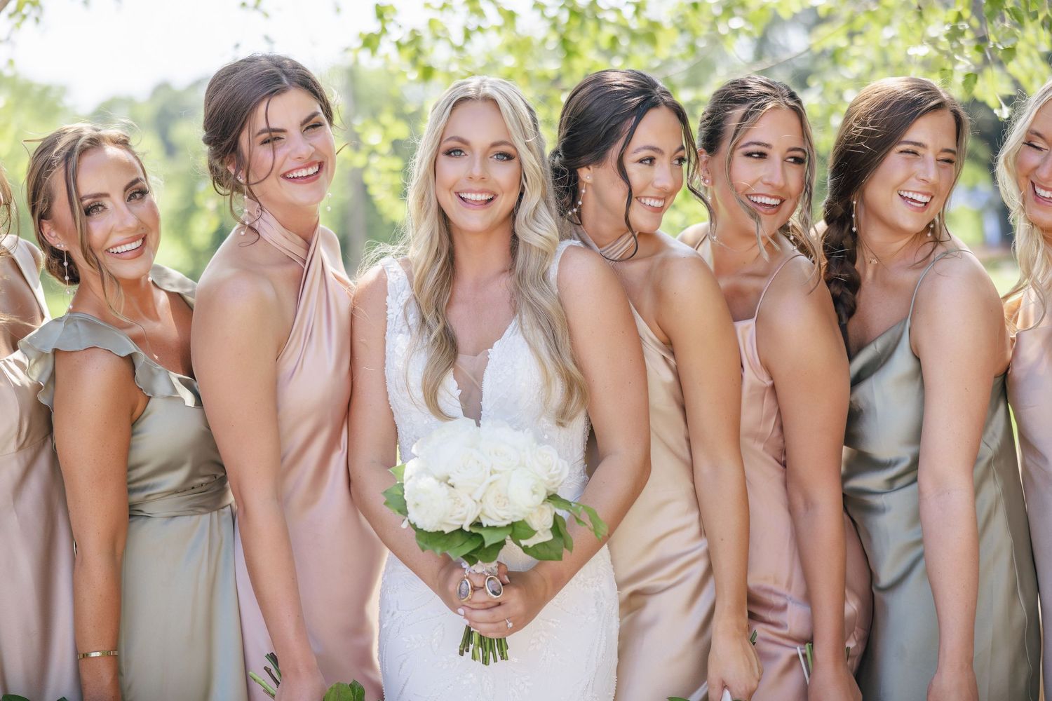 Bridal party in neutral-toned dresses pose together outdoors with bright smiles.