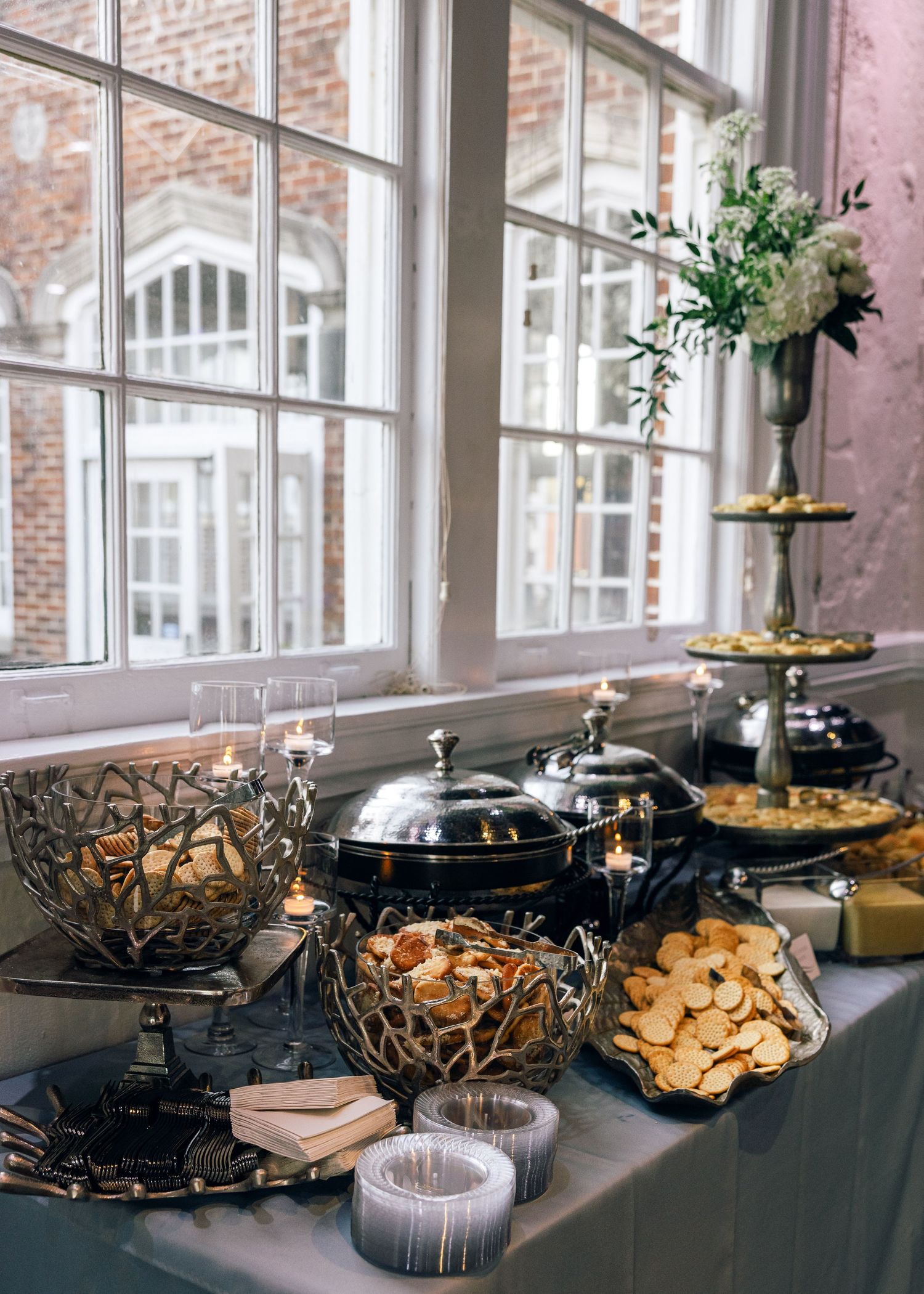 Elegant dessert and appetizer buffet display with ornate serving dishes by a bright window.