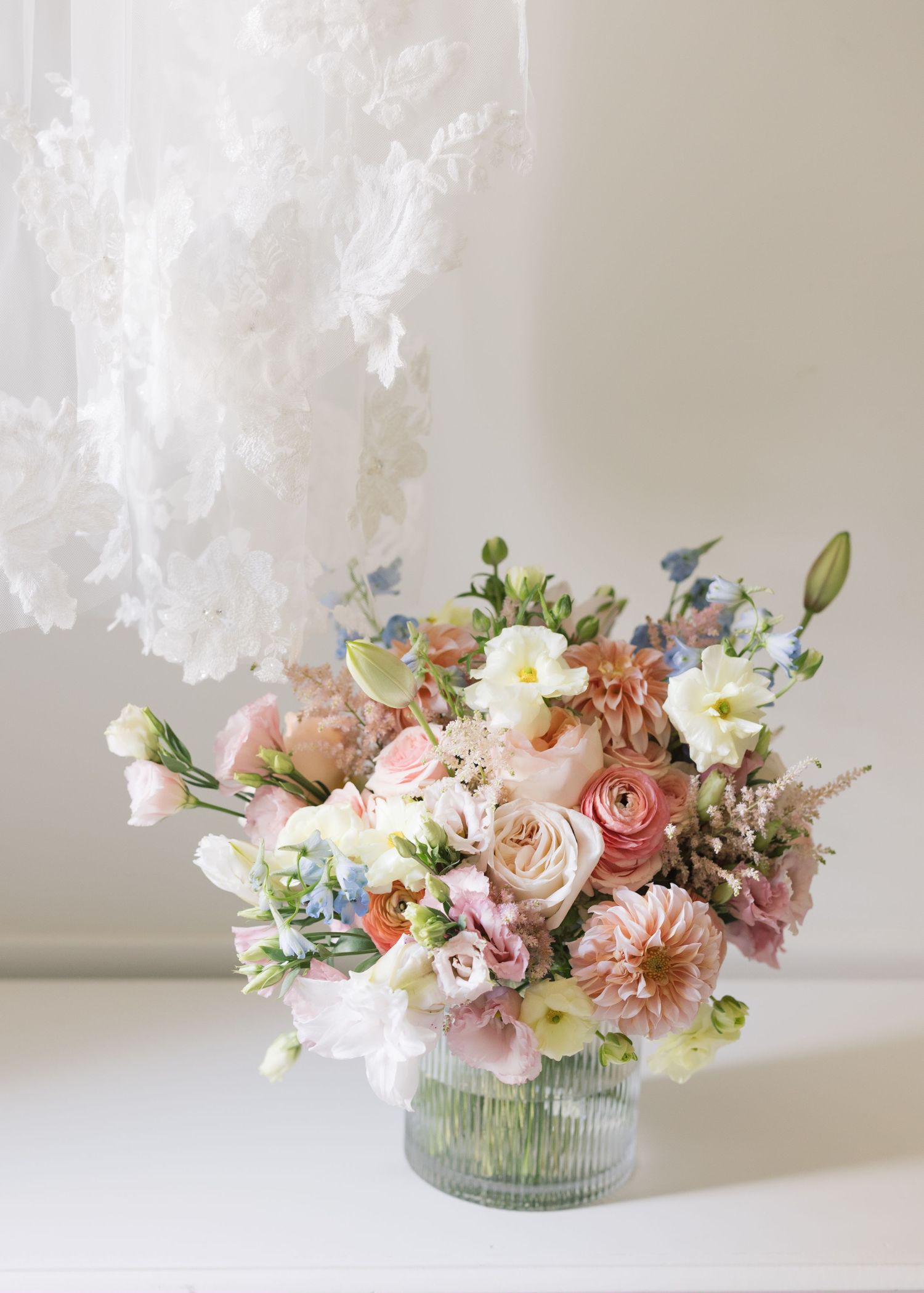 Delicate floral arrangement featuring pink roses, white blooms and blue accents in a ribbed glass vase on white surface.