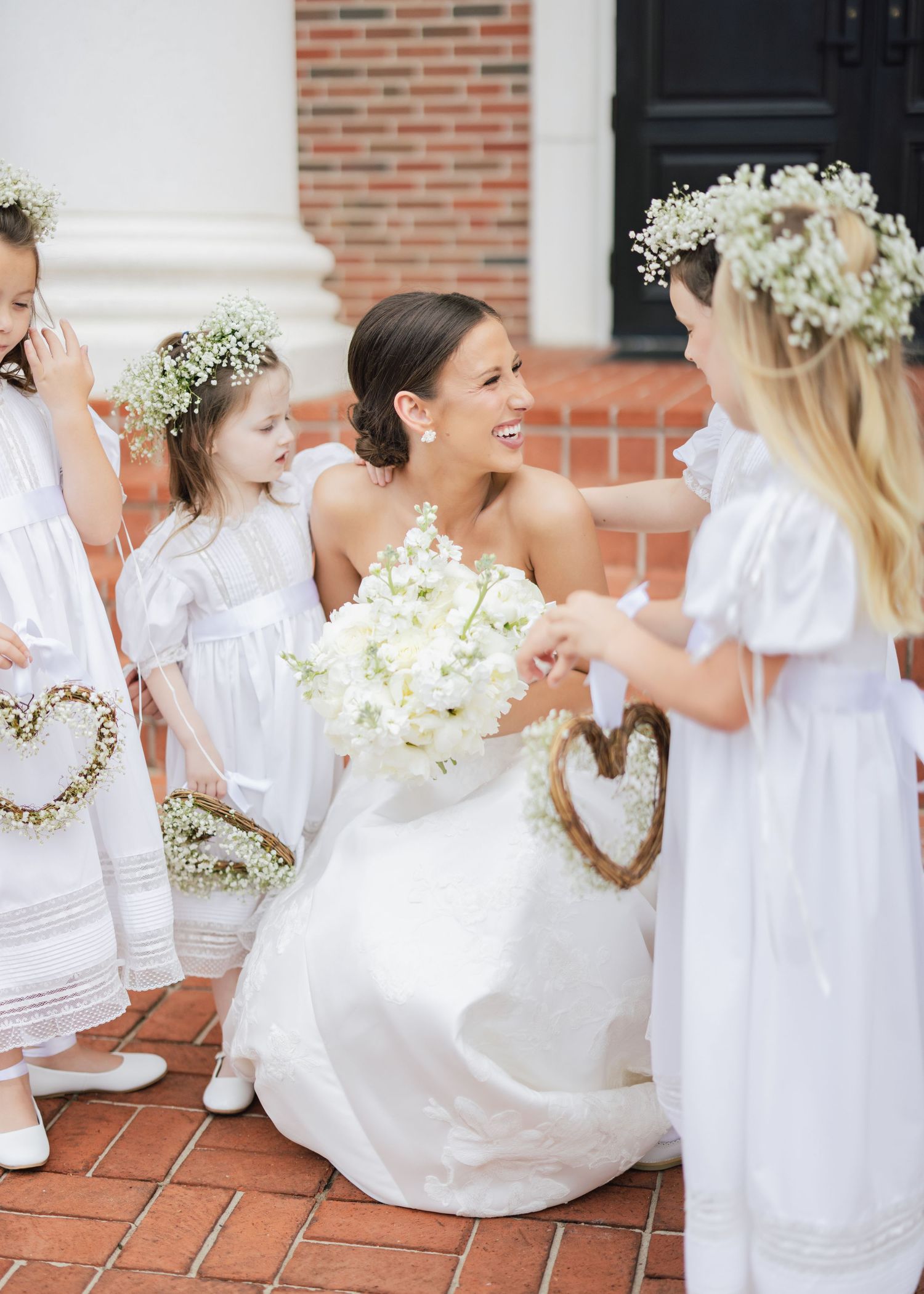 Young flower girls in white dresses sitting on brick steps with floral arrangements.
