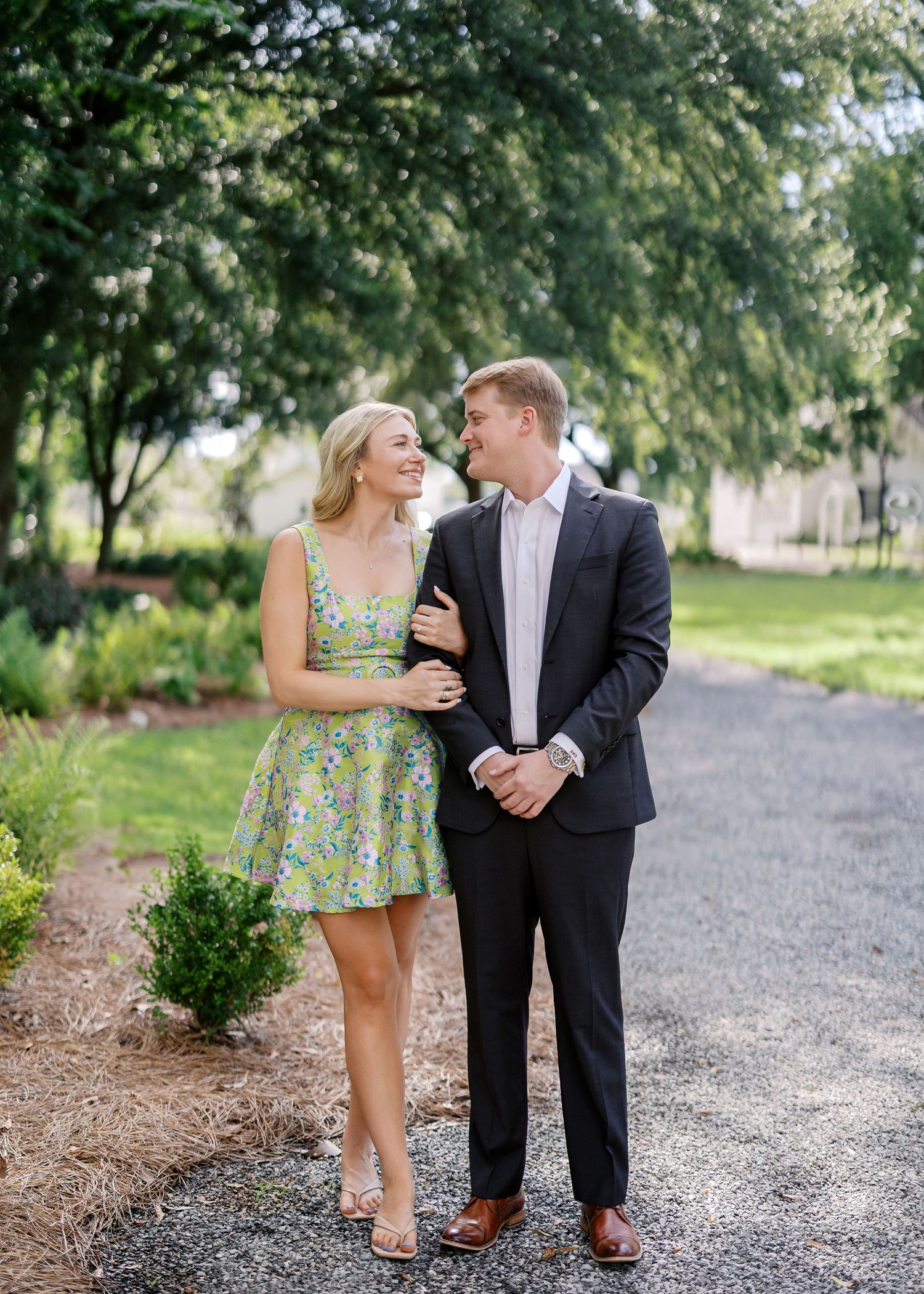 Couple in dressy attire walking along garden path under tree canopy.
