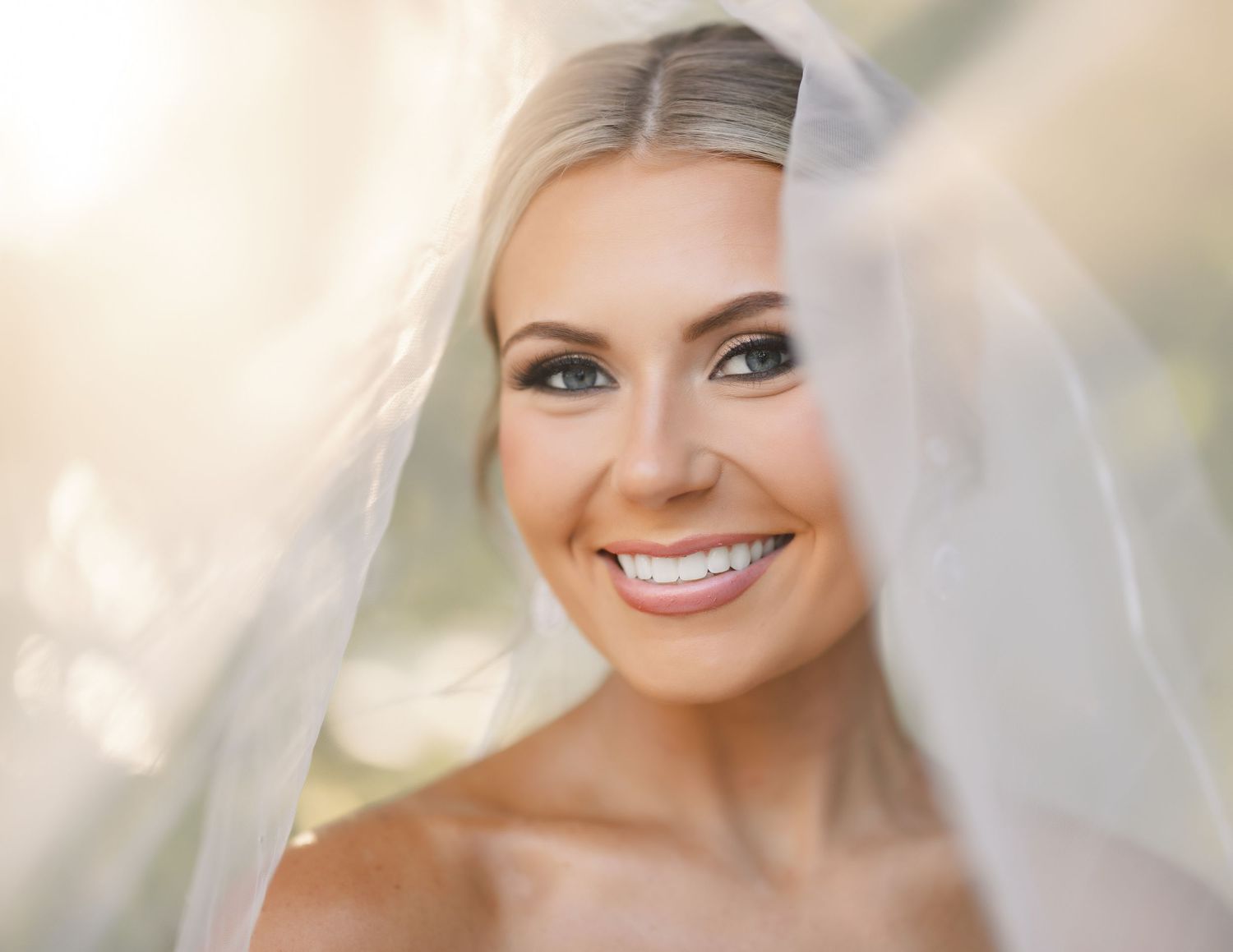 Bride wearing a delicate white veil and elegant makeup smiles radiantly during her wedding day photo session.