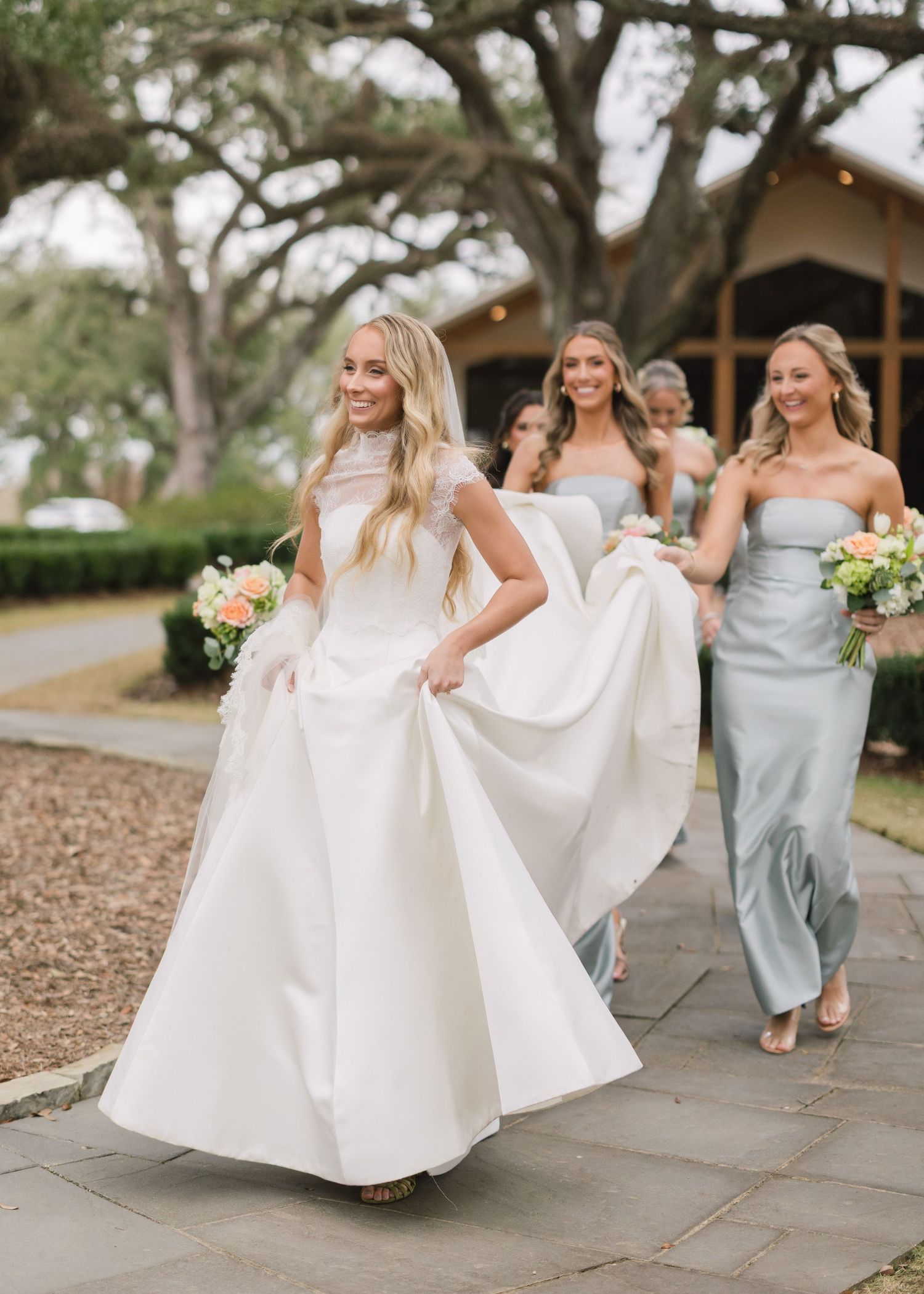 Wedding party members in sage green dresses and white gowns walk together on a stone path during an outdoor celebration.