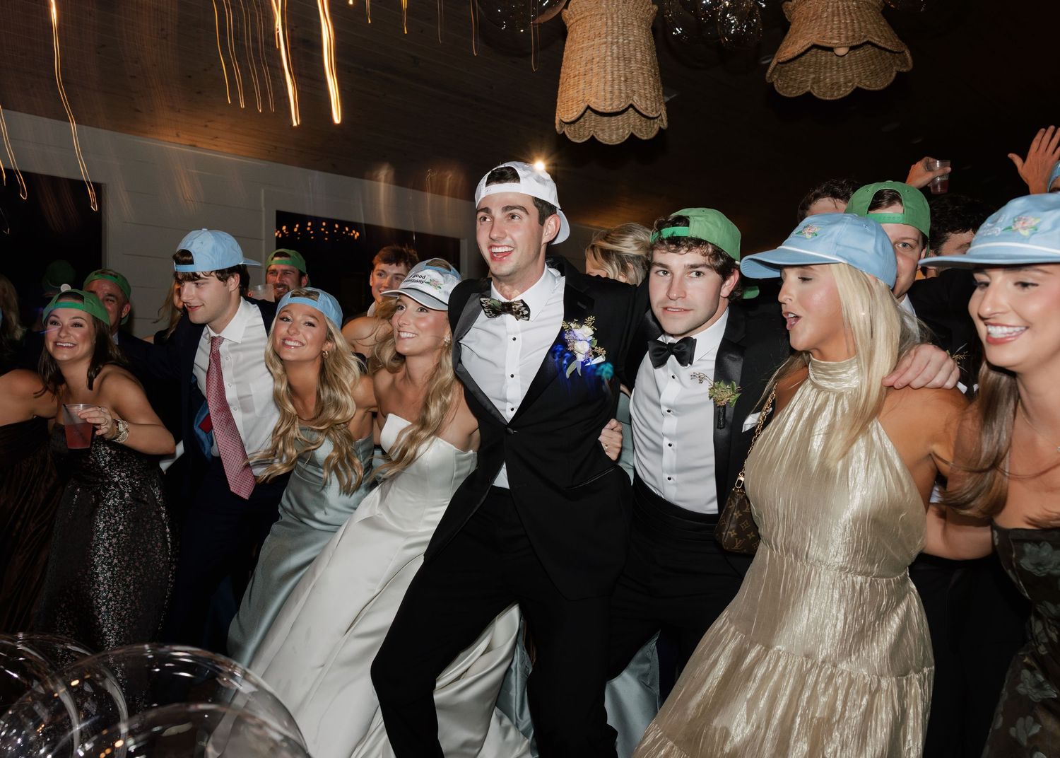 Wedding reception guests dance energetically on a wooden dance floor under twinkling lights at an indoor venue.
