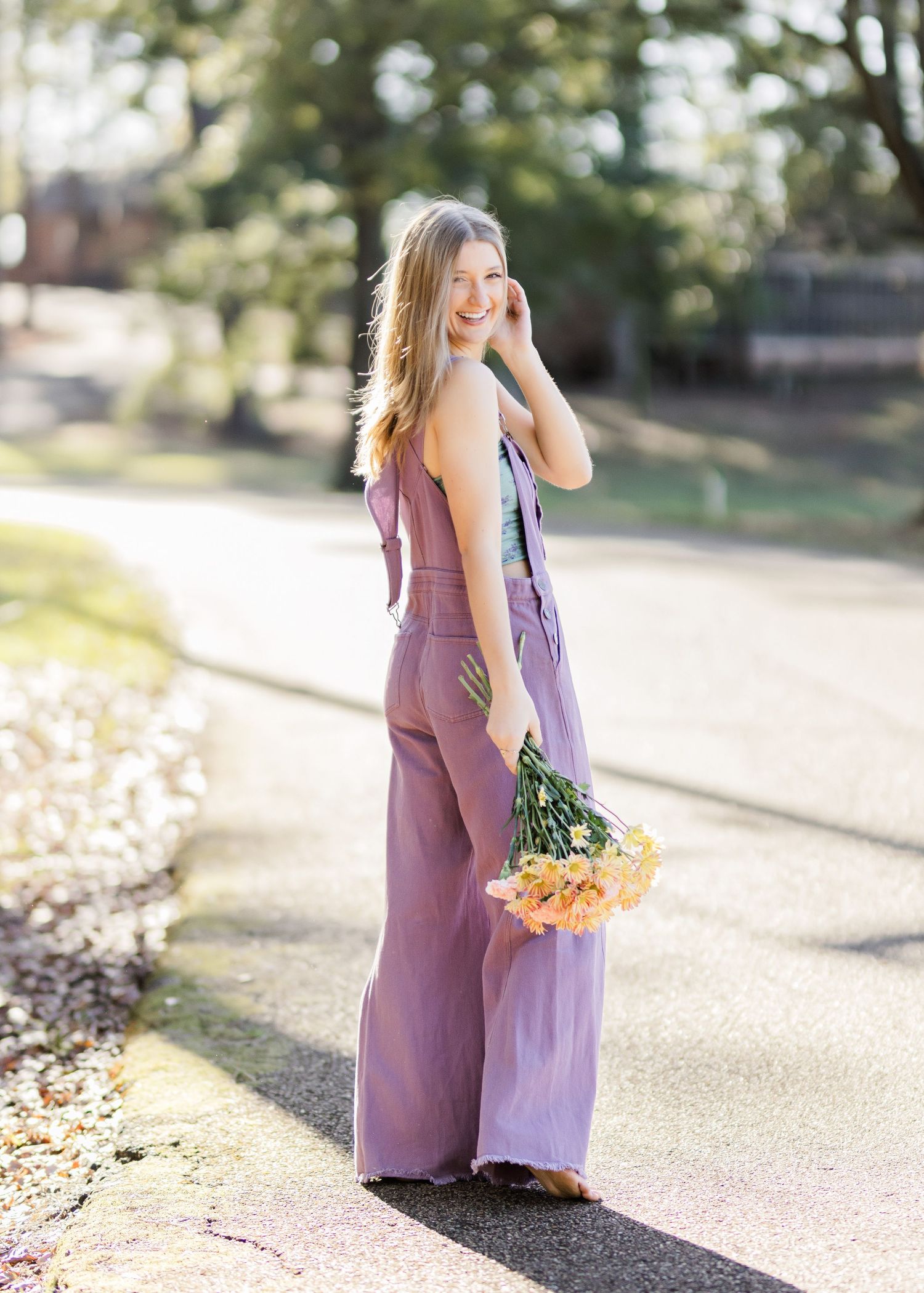A person in a mauve jumpsuit walks along a sunlit path holding orange flowers and a purple tote bag.