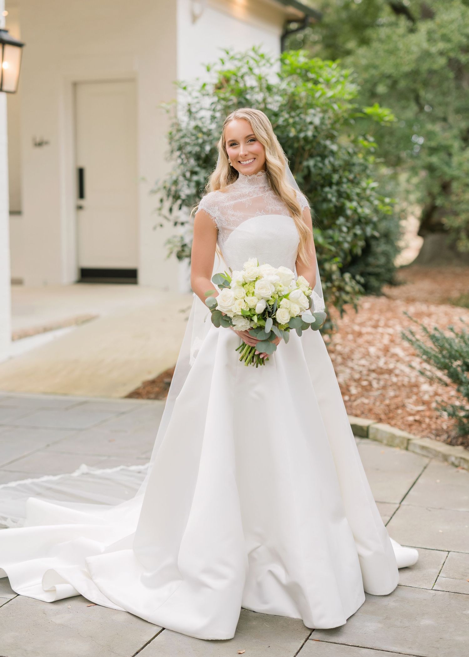 A bride in a classic white ballgown holds a bouquet while posing on stone steps surrounded by greenery.