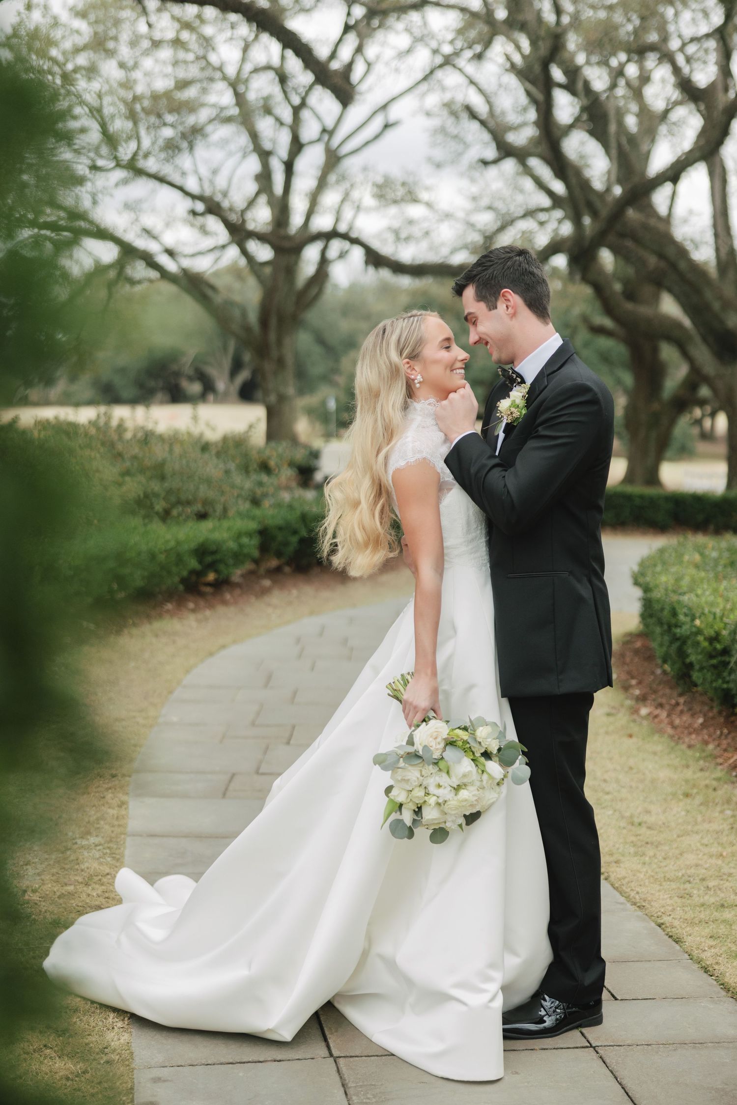 A couple in wedding attire share an intimate moment on a garden path surrounded by trees.