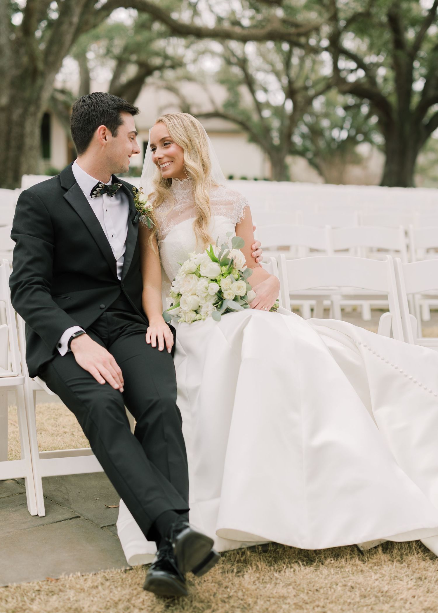 Romantic wedding couple shares intimate moments while sitting on white chairs at an outdoor garden ceremony venue.