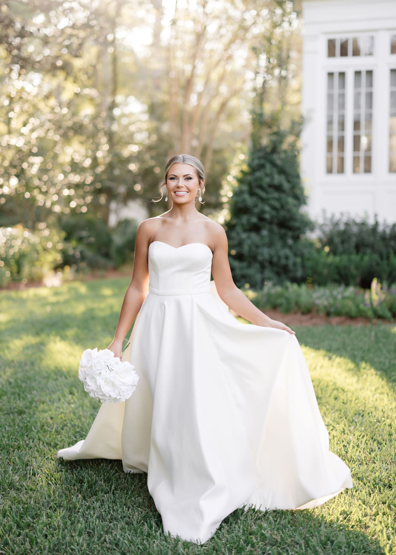 A bride in a flowing white wedding gown sits gracefully on lush green grass during a golden sunset.