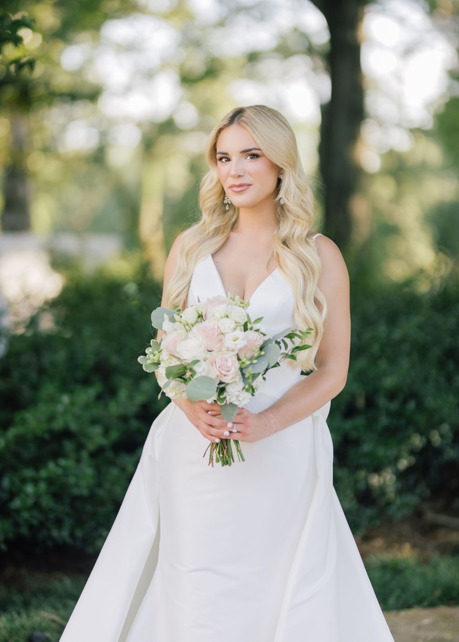 Bride in white dress holding delicate pastel bouquet in outdoor garden setting.
