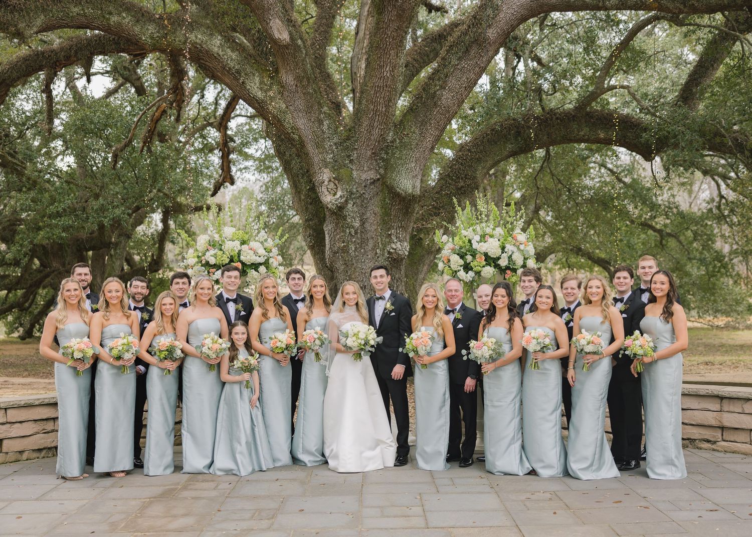 Wedding party in light blue dresses and dark suits poses together under large oak trees in outdoor setting.