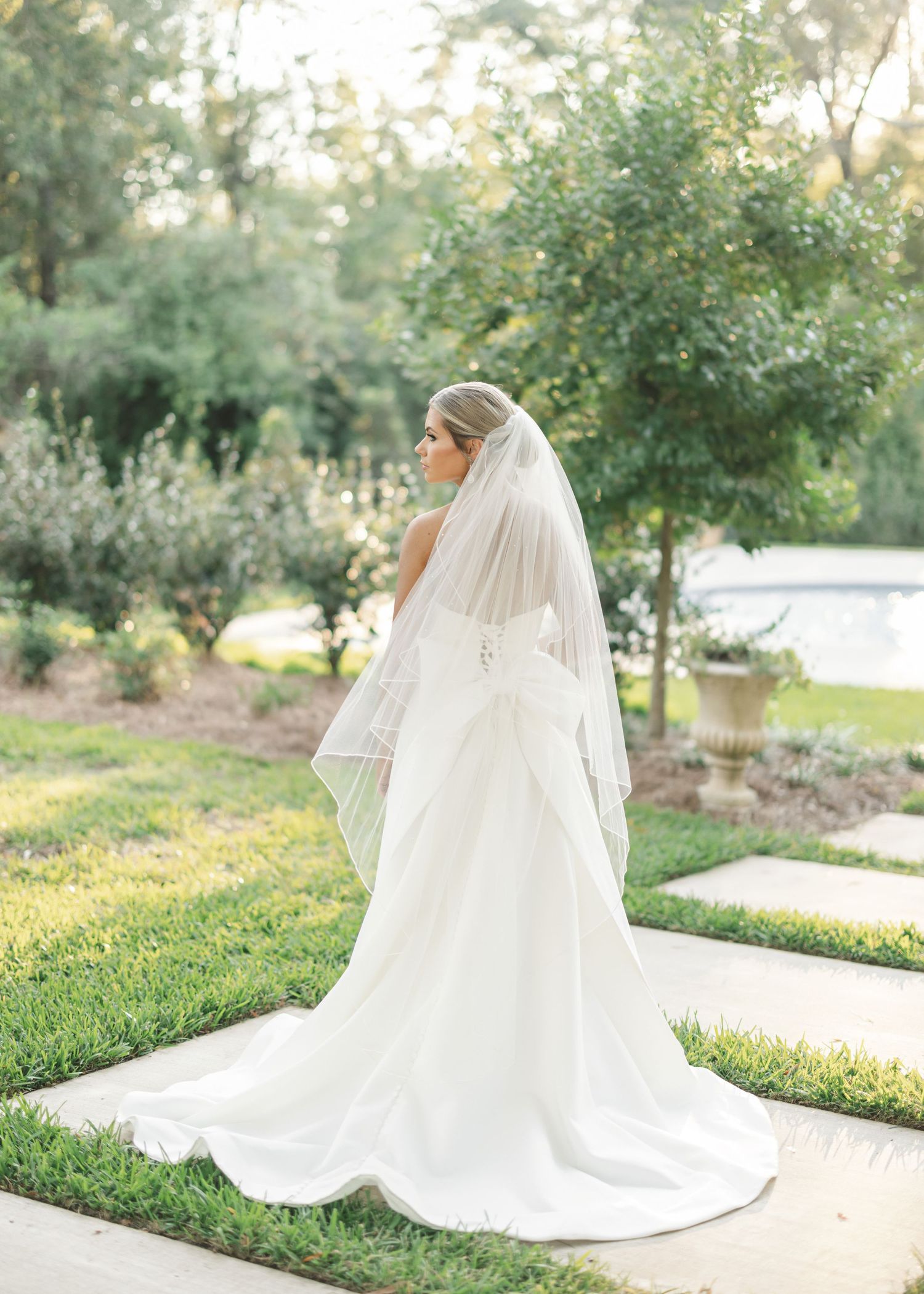 A flowing white wedding dress with a dramatic cathedral-length veil photographed in a garden setting at sunset.