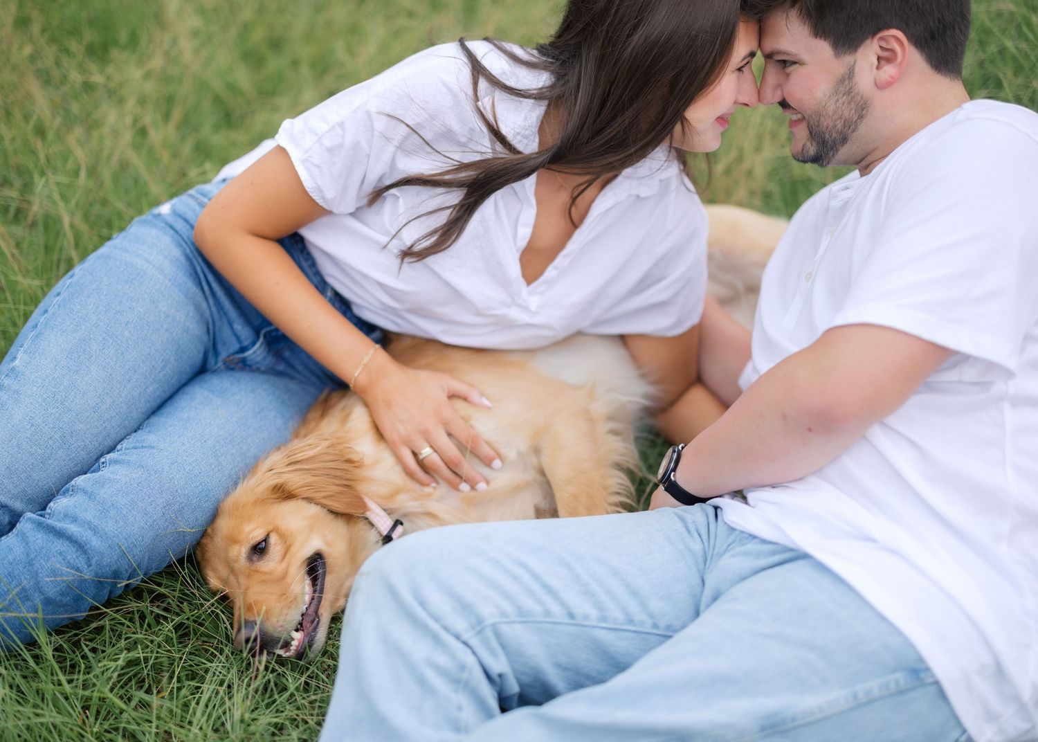 A couple in casual wear petting their golden retriever while sitting in grass.