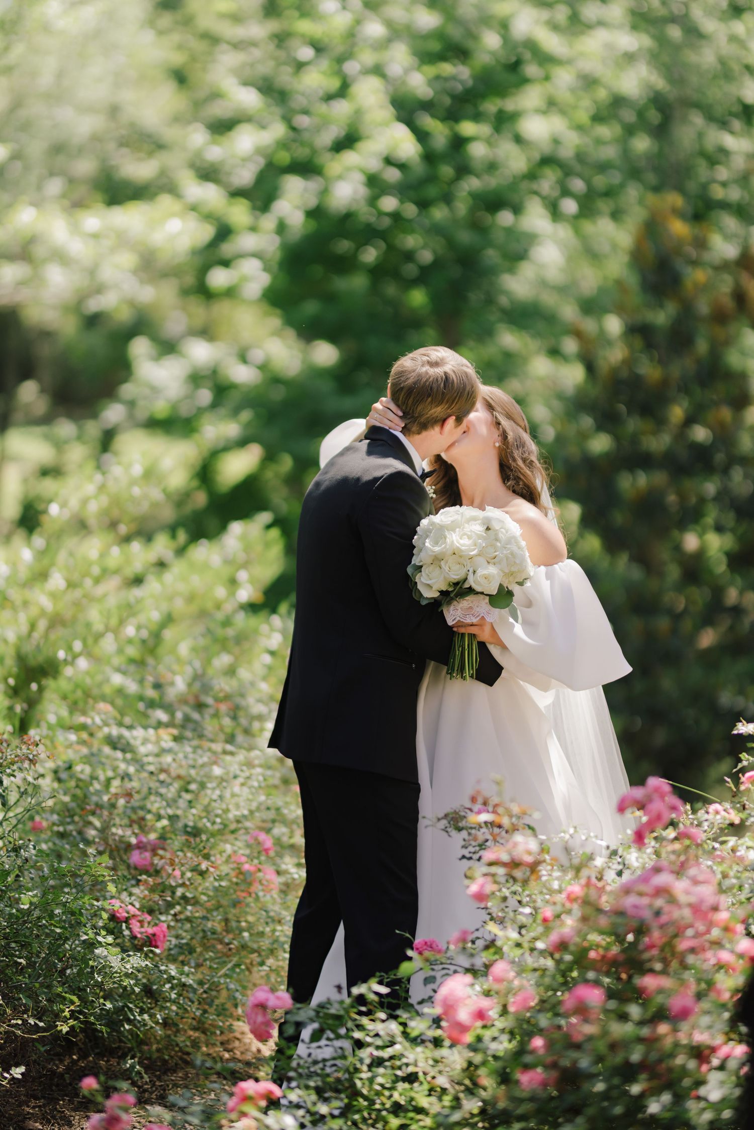 Romantic wedding couple sharing a kiss in a blooming garden with pink flowers.
