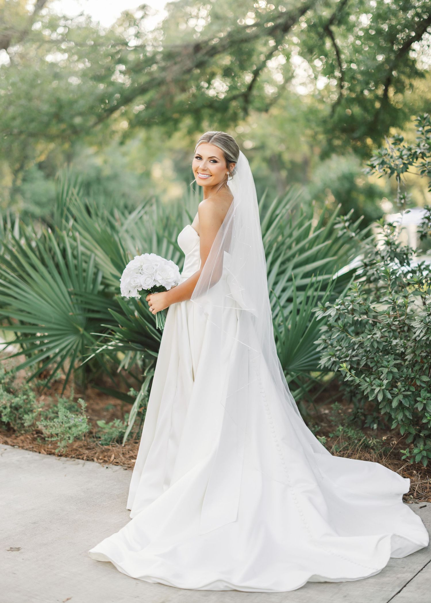 Bride in flowing white wedding gown walks through a tropical garden path with palm fronds.