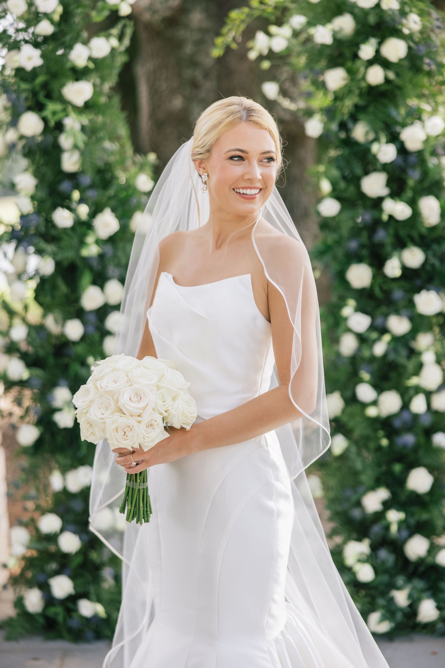 A bride in a white sleeveless gown holds a bouquet against a backdrop of white roses.
