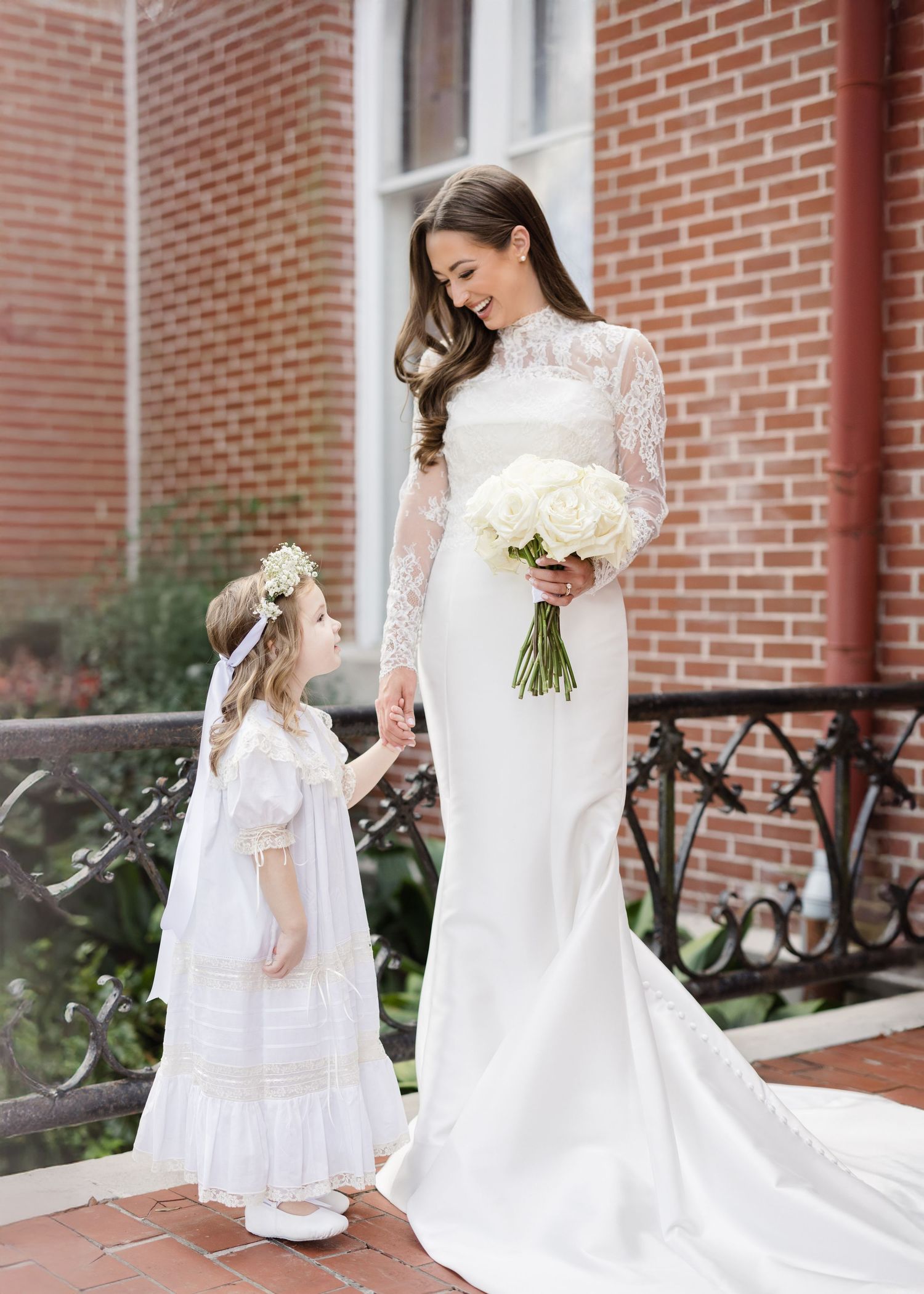 Adorable flower girl in white dress walks with bride in elegant wedding gown against brick wall.