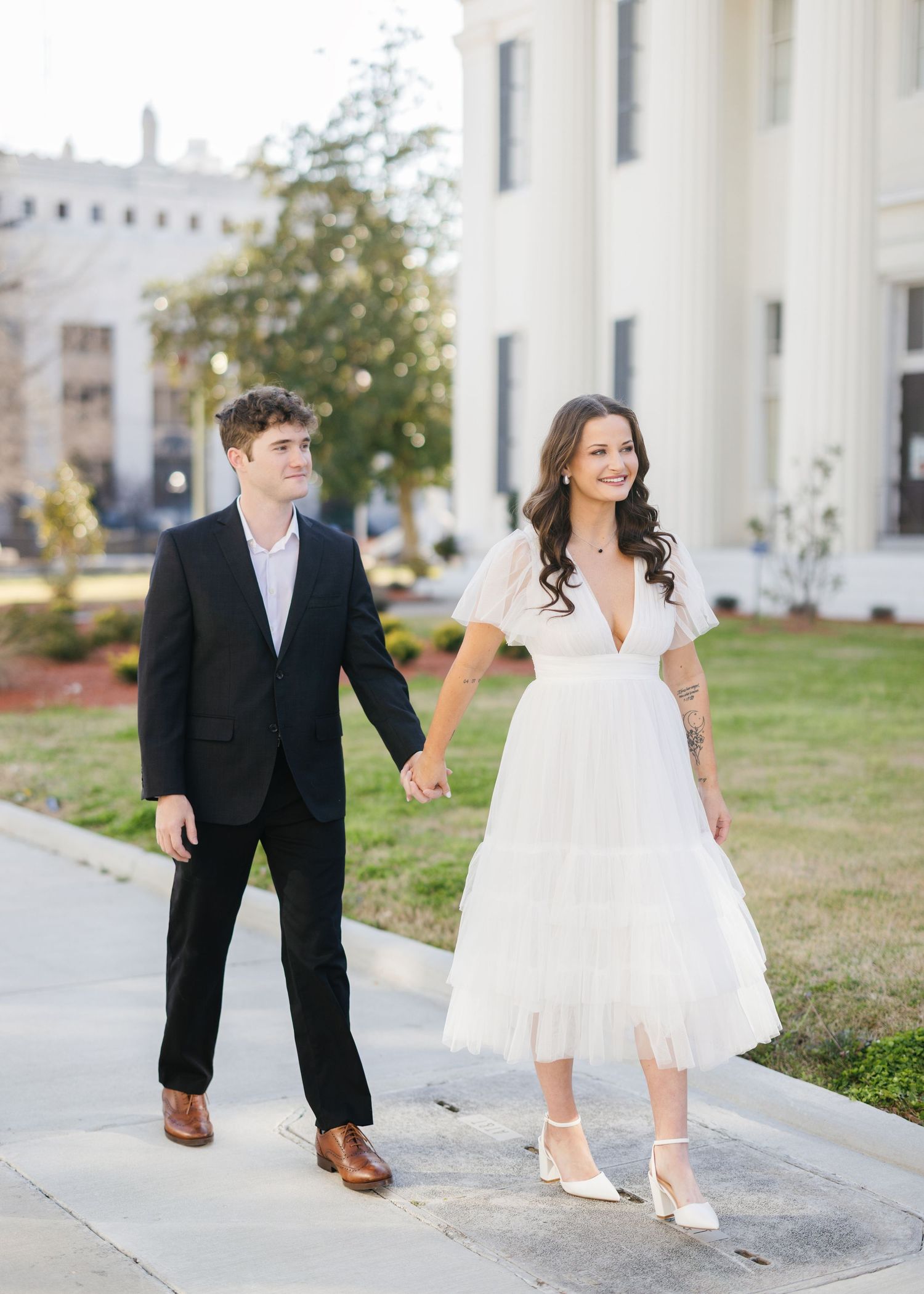 Playful couple walks along white building pathway in formal attire.