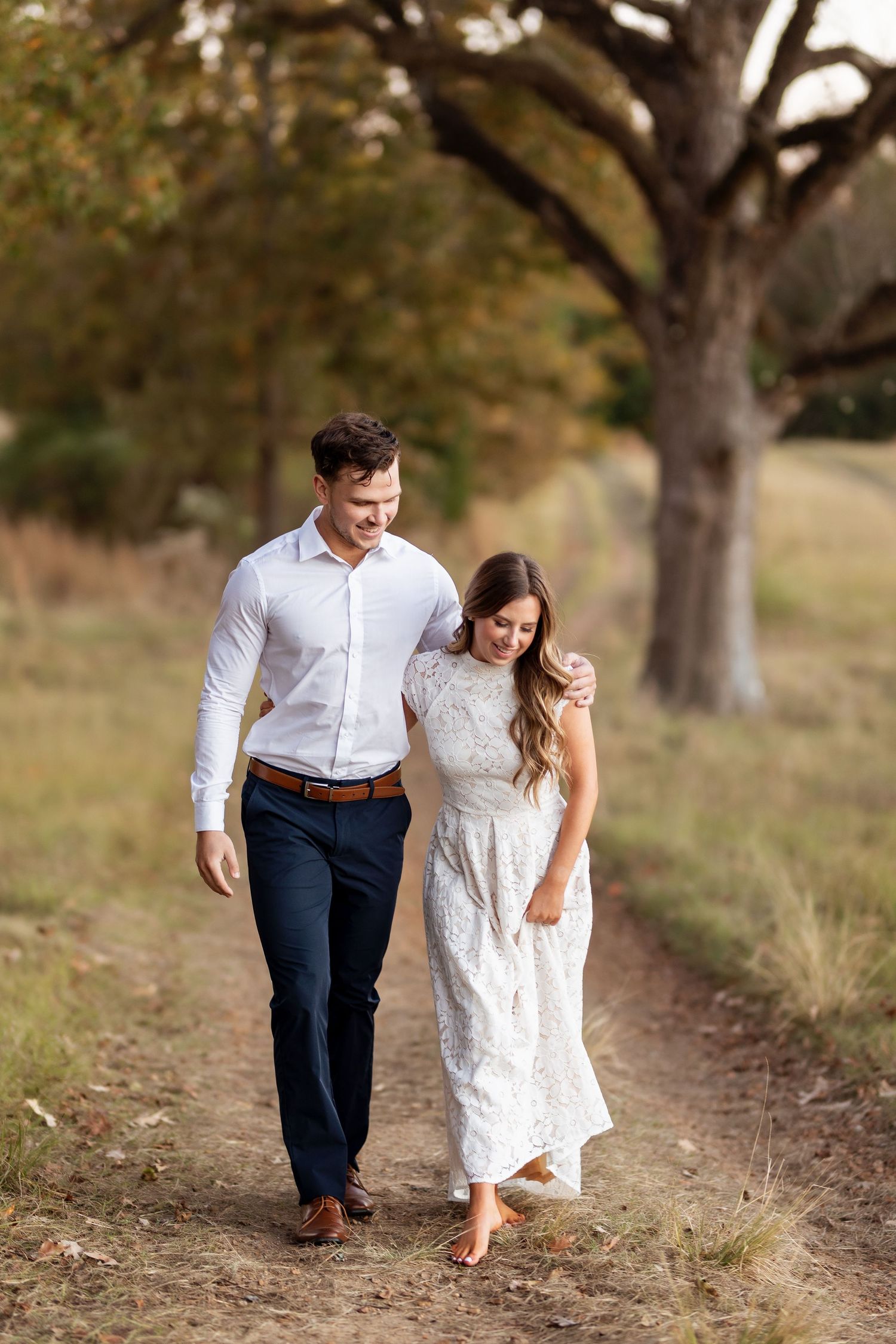 A couple walks hand in hand down a tree-lined dirt path in casual wedding attire.