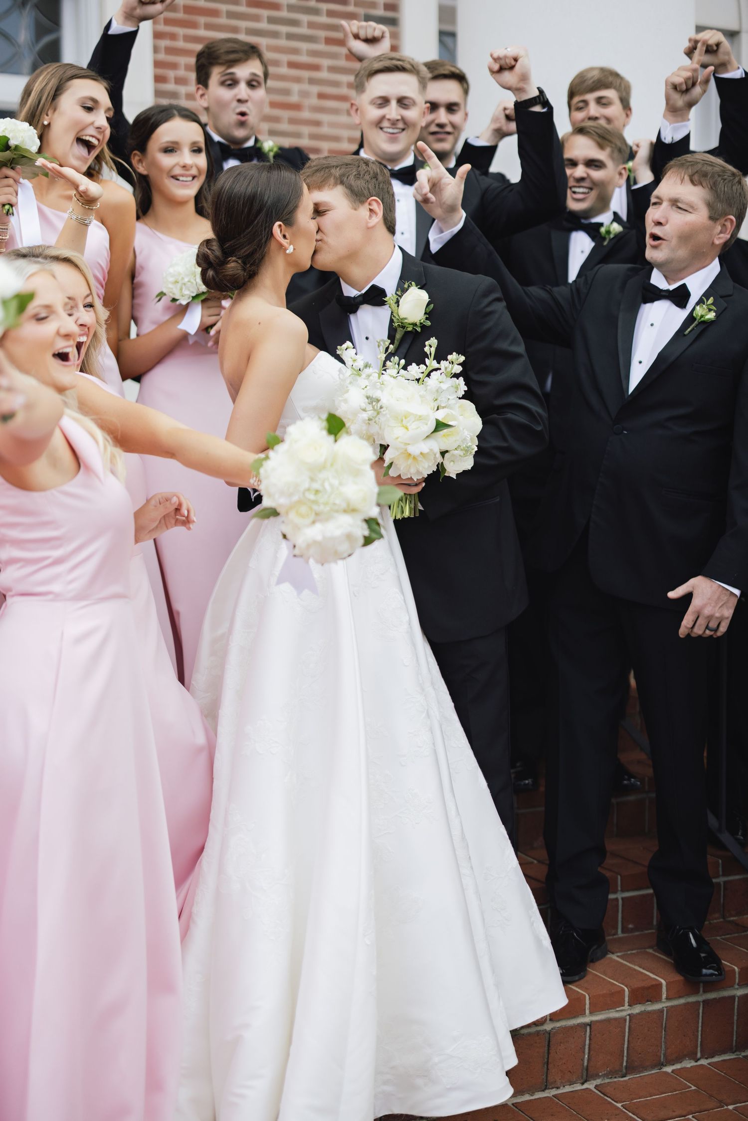 Wedding party poses together wearing pink bridesmaid dresses and black tuxedos.