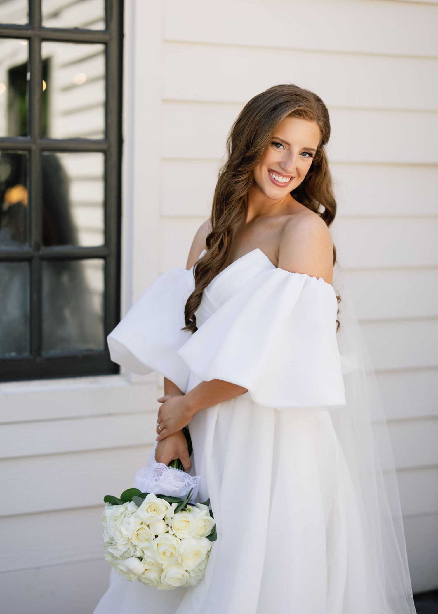 A bride in an off-shoulder white gown holds a bouquet of white flowers against a white exterior wall.
