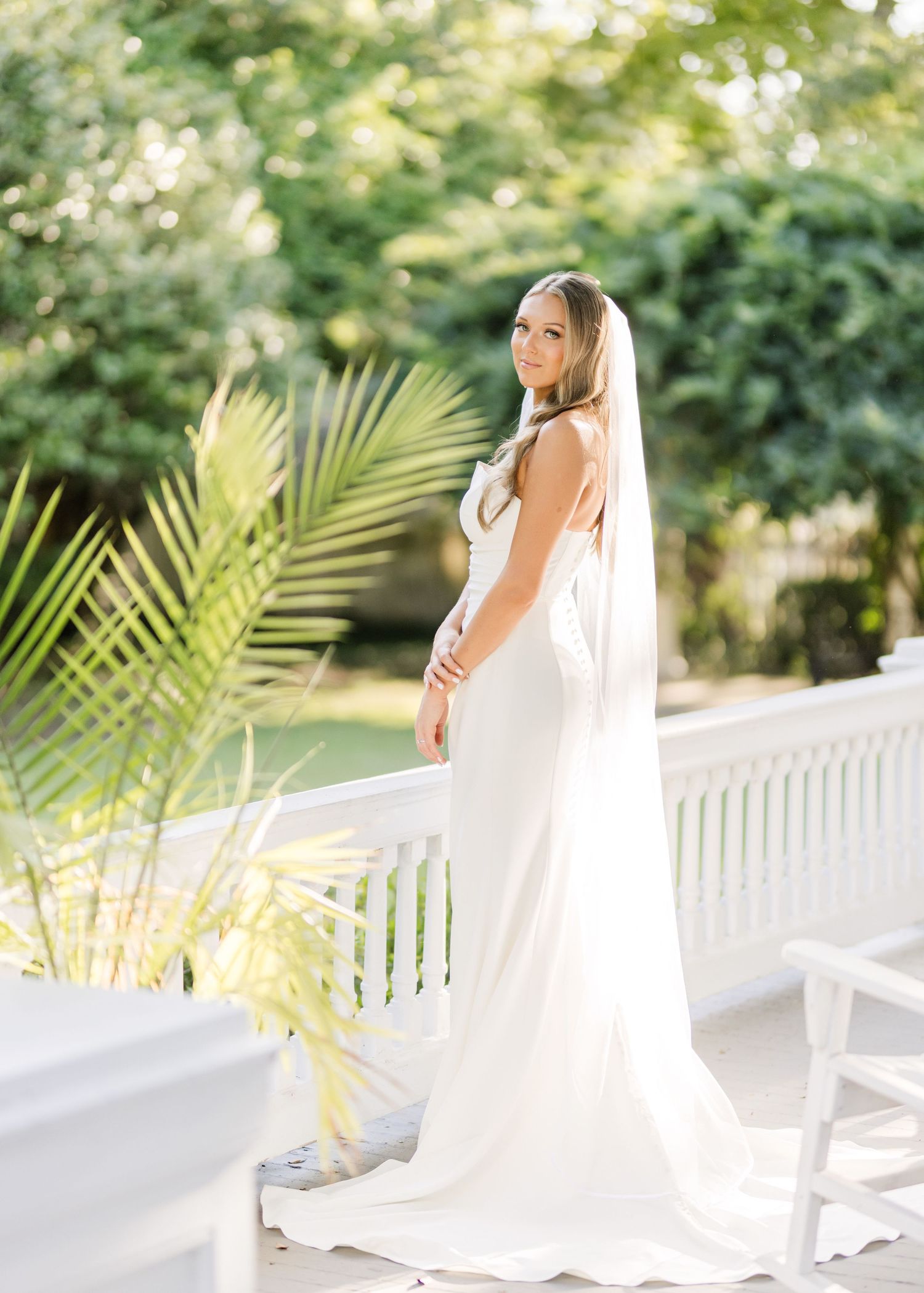 Bride in elegant white gown poses by tropical palms and white railing.