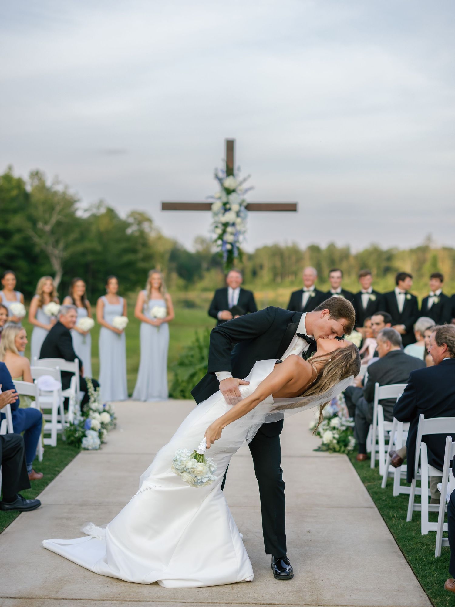 Romantic wedding couple performs a dip kiss on a paved path lined with white chairs during an outdoor ceremony.