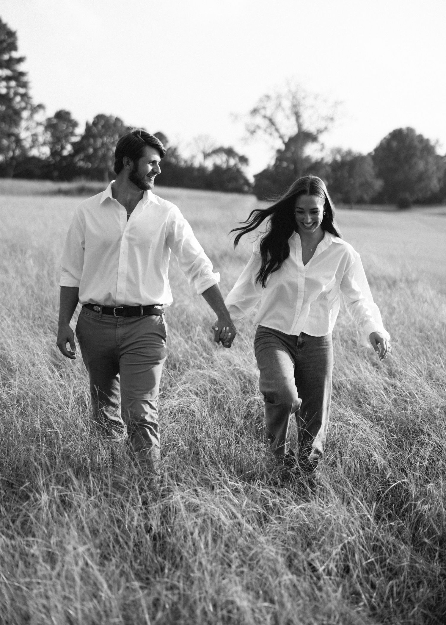 Black and white photo of couple walking hand in hand through tall grass field.