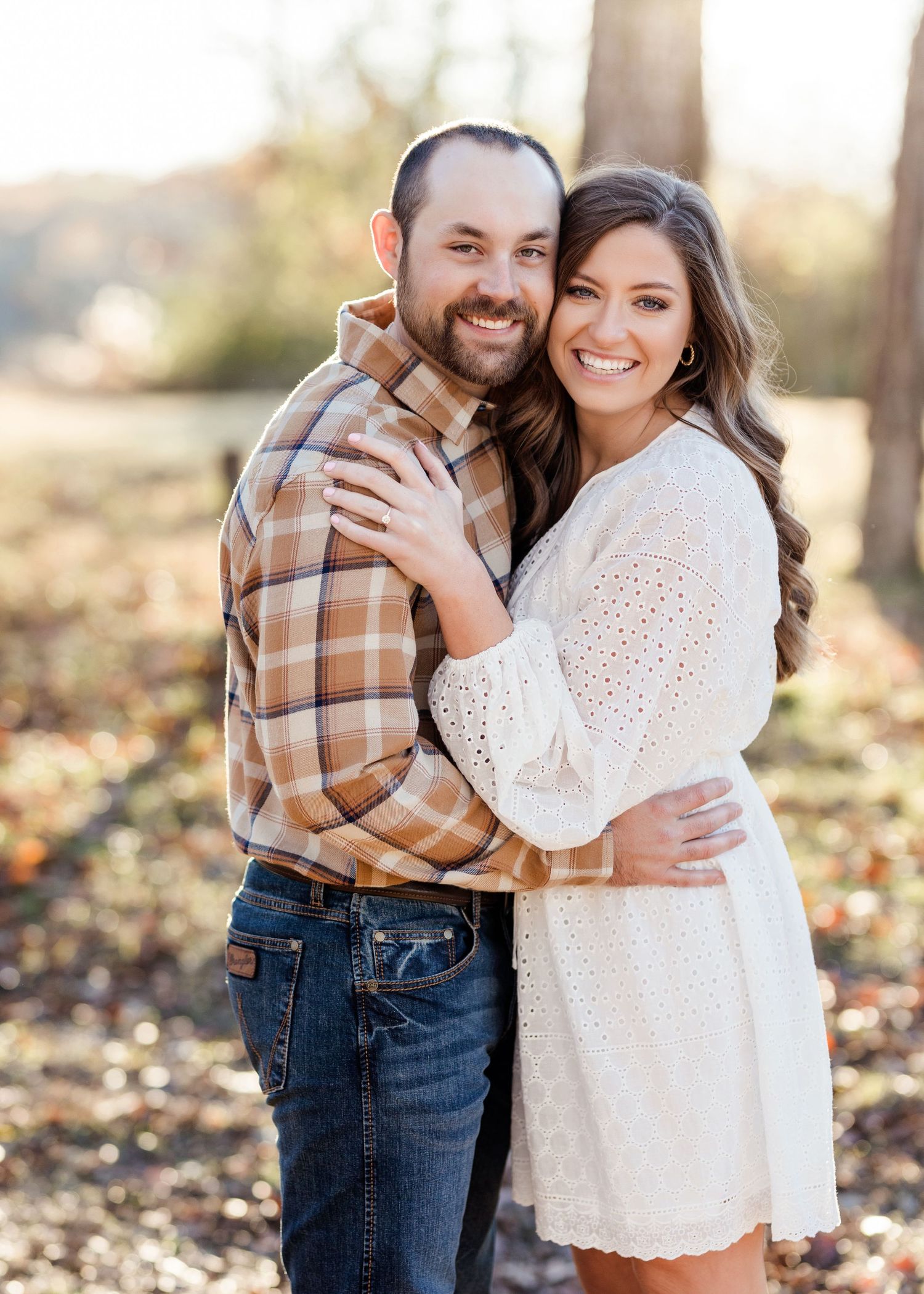 Couple embracing outdoors in casual attire with woman in white dress and man in plaid shirt.