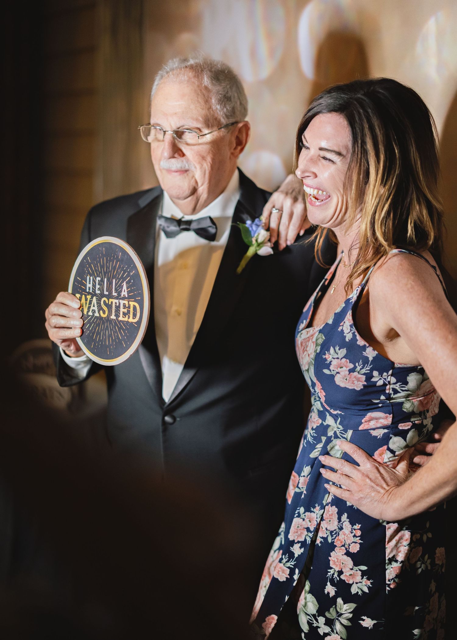 Two wedding guests share a laugh while holding a novelty 'Bella Wasted' sign at a reception celebration.
