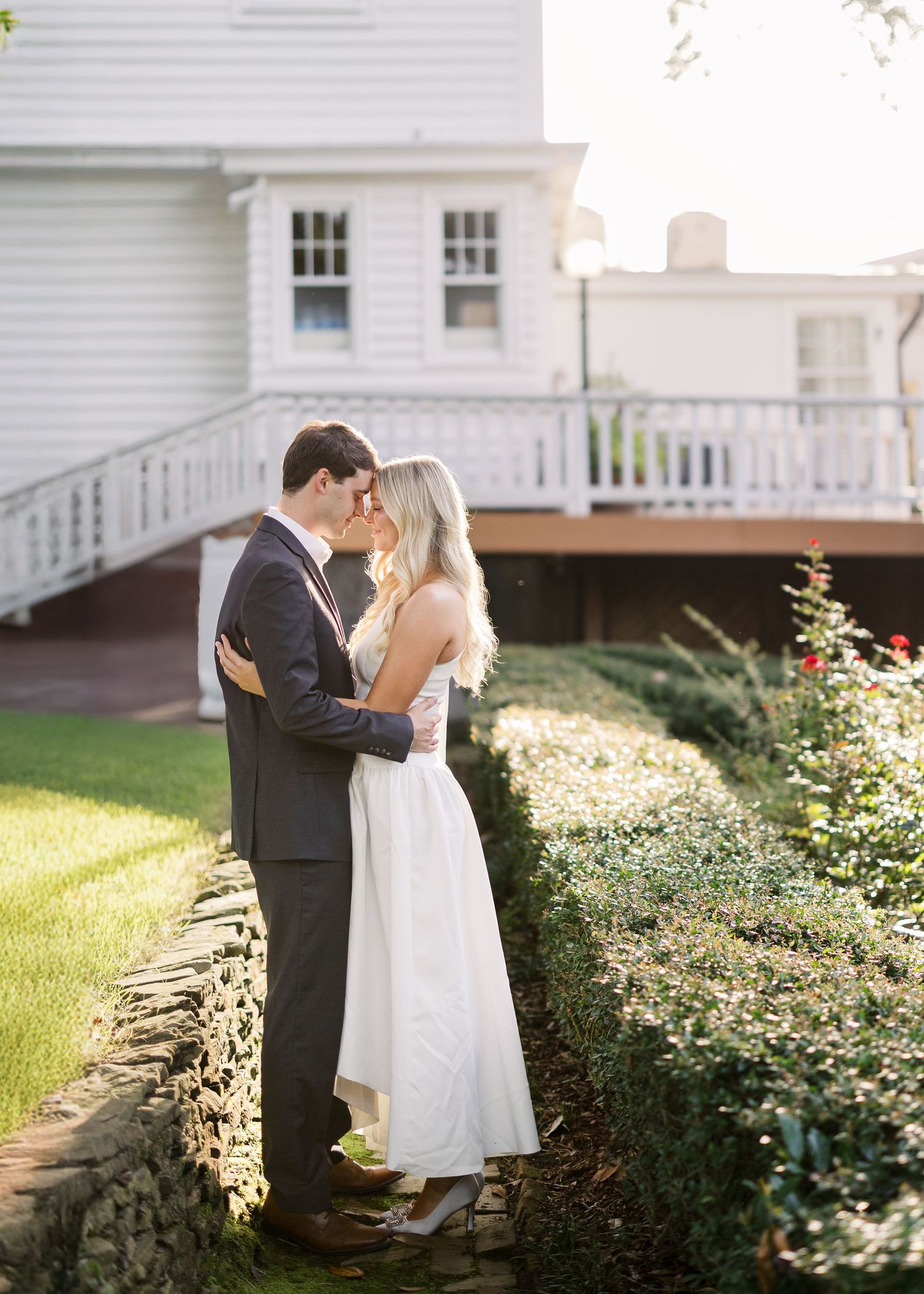 Romantic moment captured in garden with white colonial house and stone wall backdrop.