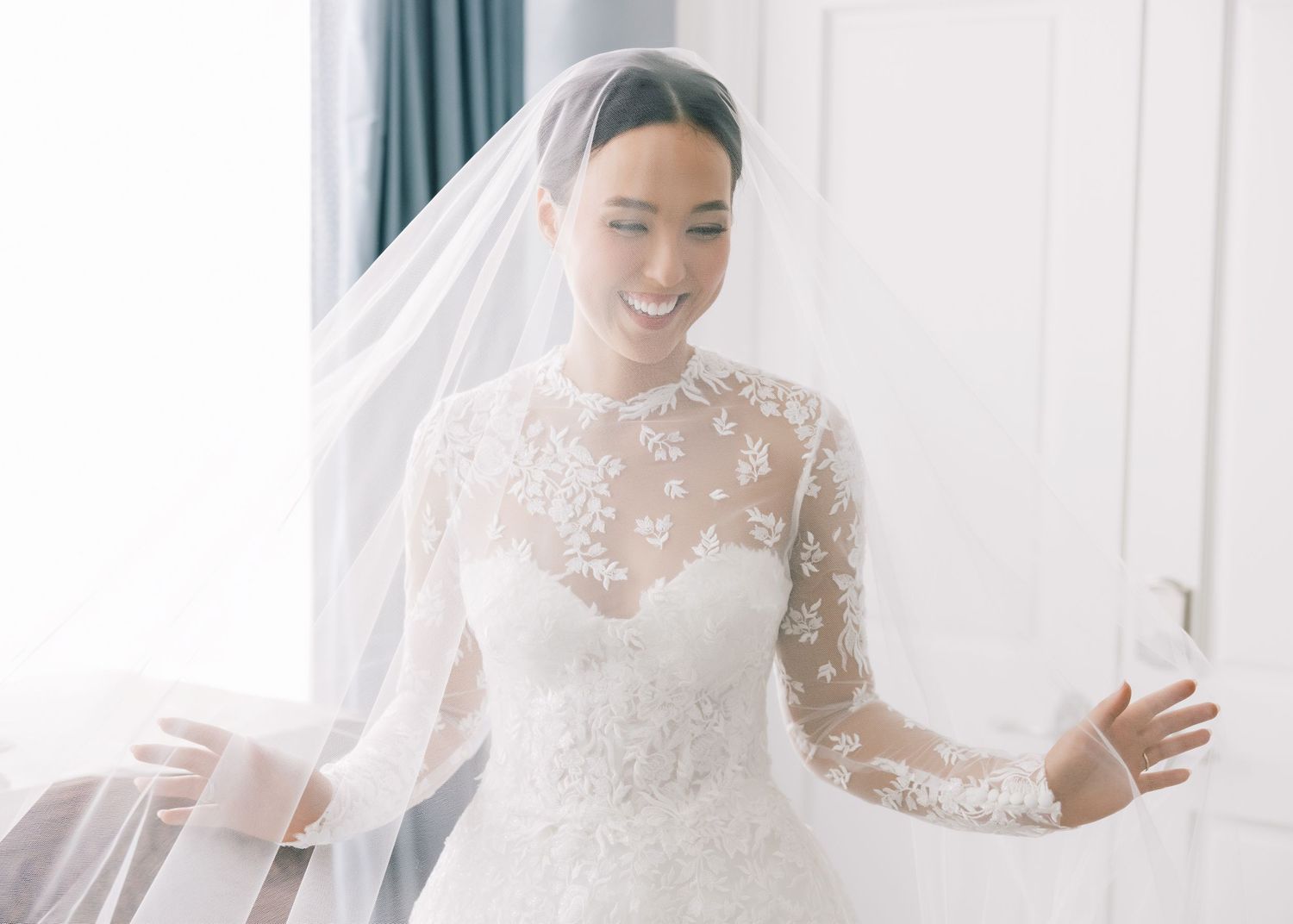 Bride in lace wedding gown poses by window with delicate veil.