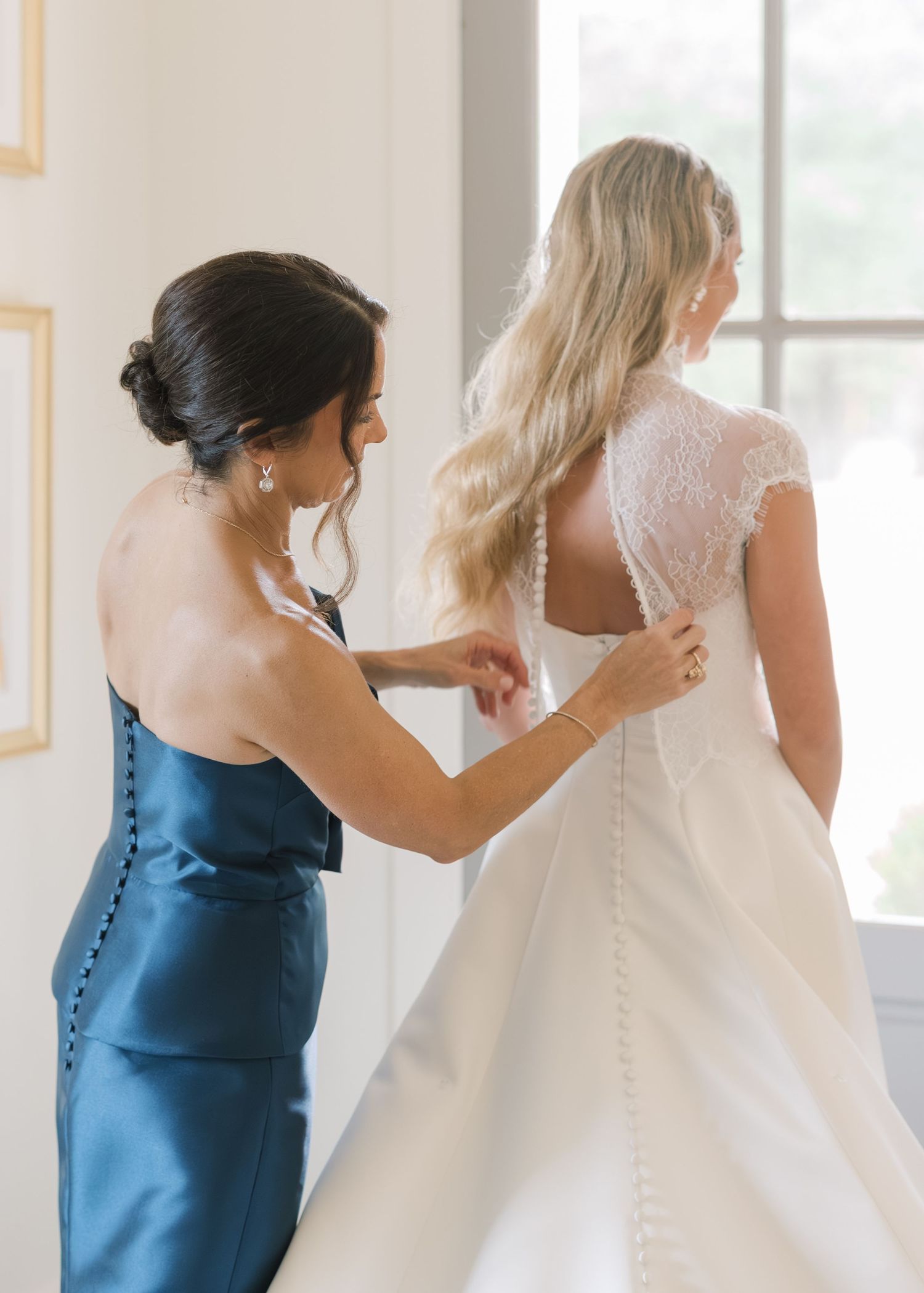 Bridesmaids in elegant blue dresses help adjust a bride's white wedding gown in a sunlit room.