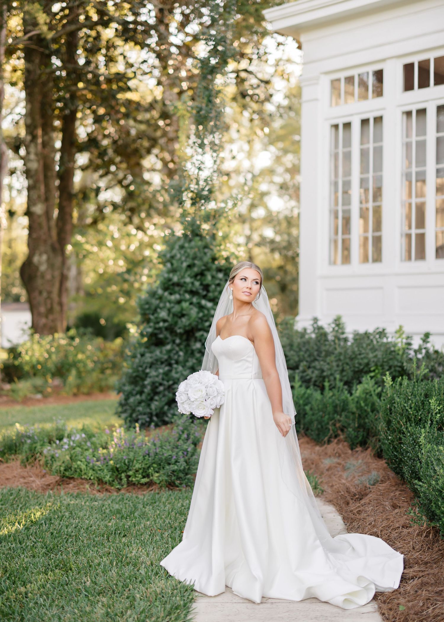 A bride in a flowing white satin wedding gown poses elegantly in a garden setting with white flowers at sunset.
