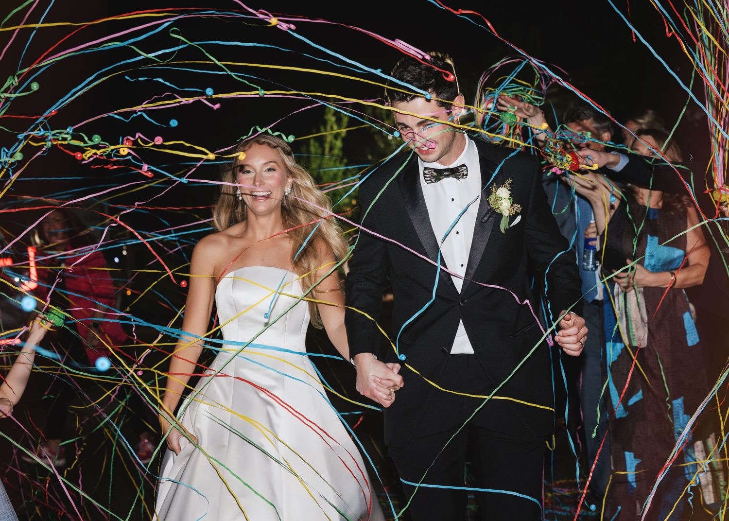 Wedding couple exits their reception through a tunnel of colorful light streamer trails at night.